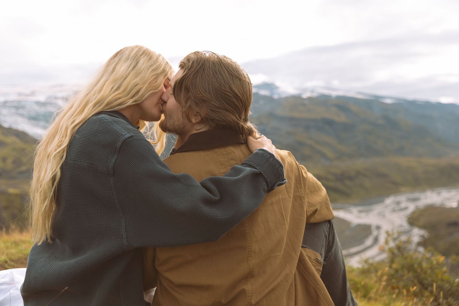 Couple sharing a kiss on a scenic mountain overlook with lush green valley and cloudy sky behind.