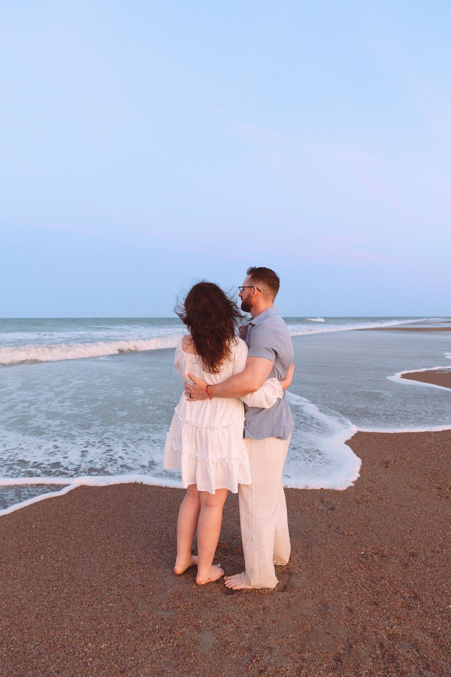 Romantic couple embracing on a beach at dusk, woman in white dress, man in gray shirt, ocean waves behind.