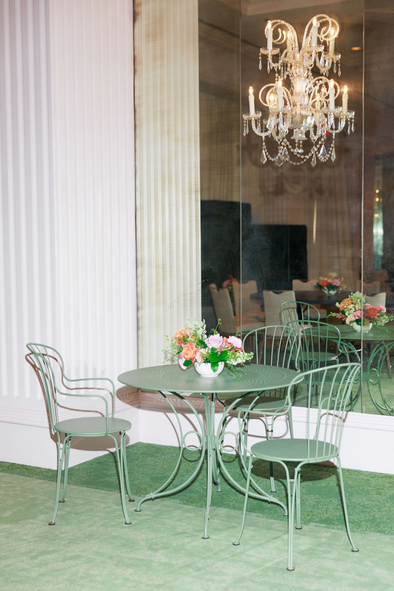 Elegant green metal bistro table and chairs set with floral centerpiece beneath a crystal chandelier.