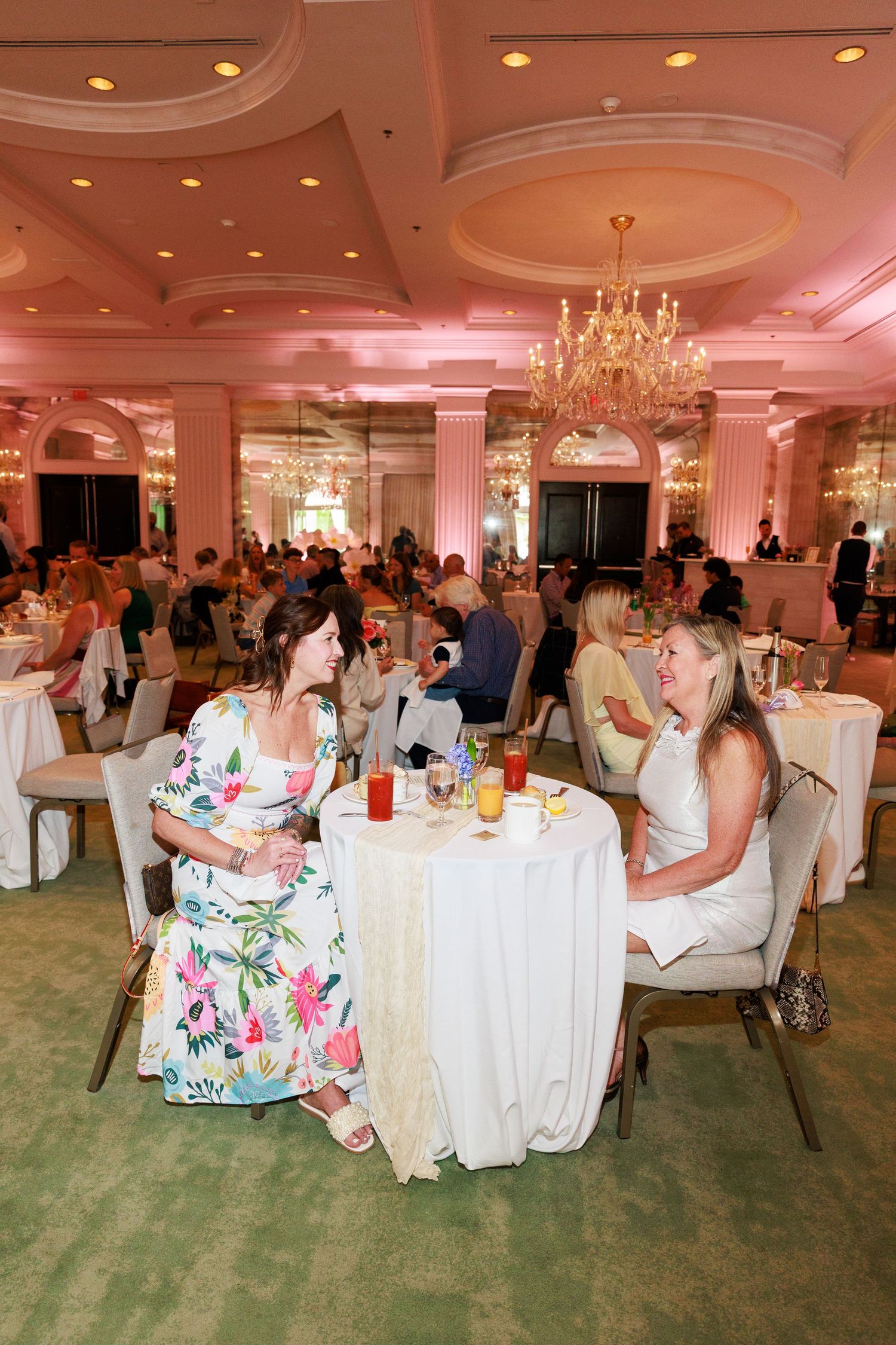Two women in elegant dresses chat over cocktails at a white-clothed table in a pink-lit ballroom.