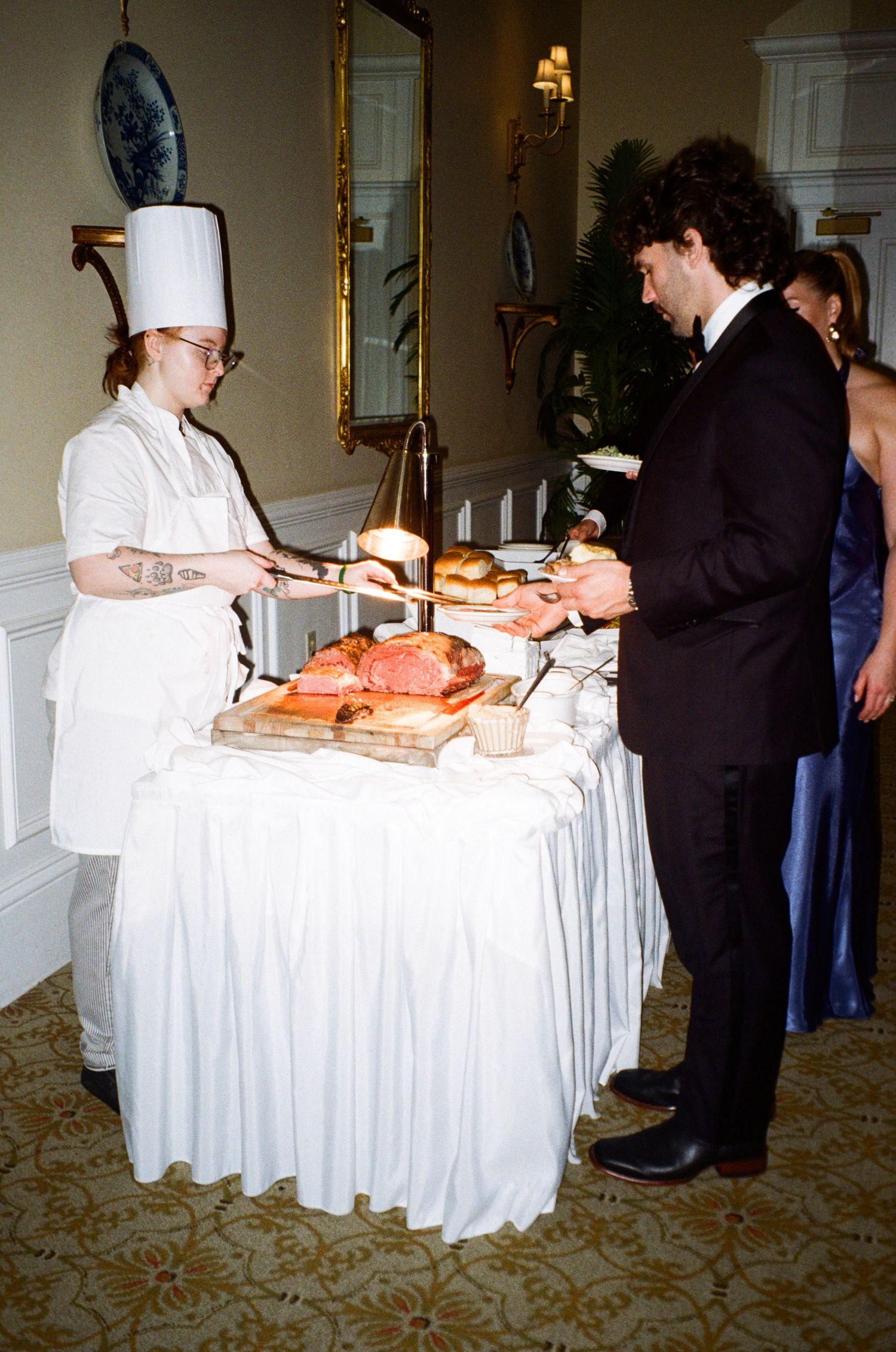 A chef in white uniform serves carved roast beef to a guest in a tuxedo at a formal catered event.