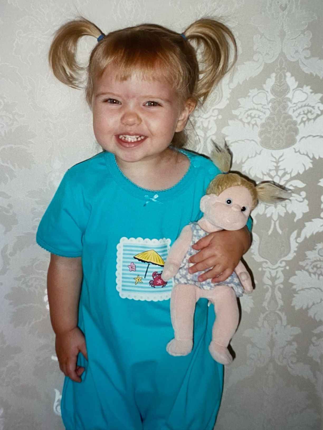 Smiling toddler girl with pigtails in teal jumpsuit holding a blonde doll with matching outfit.