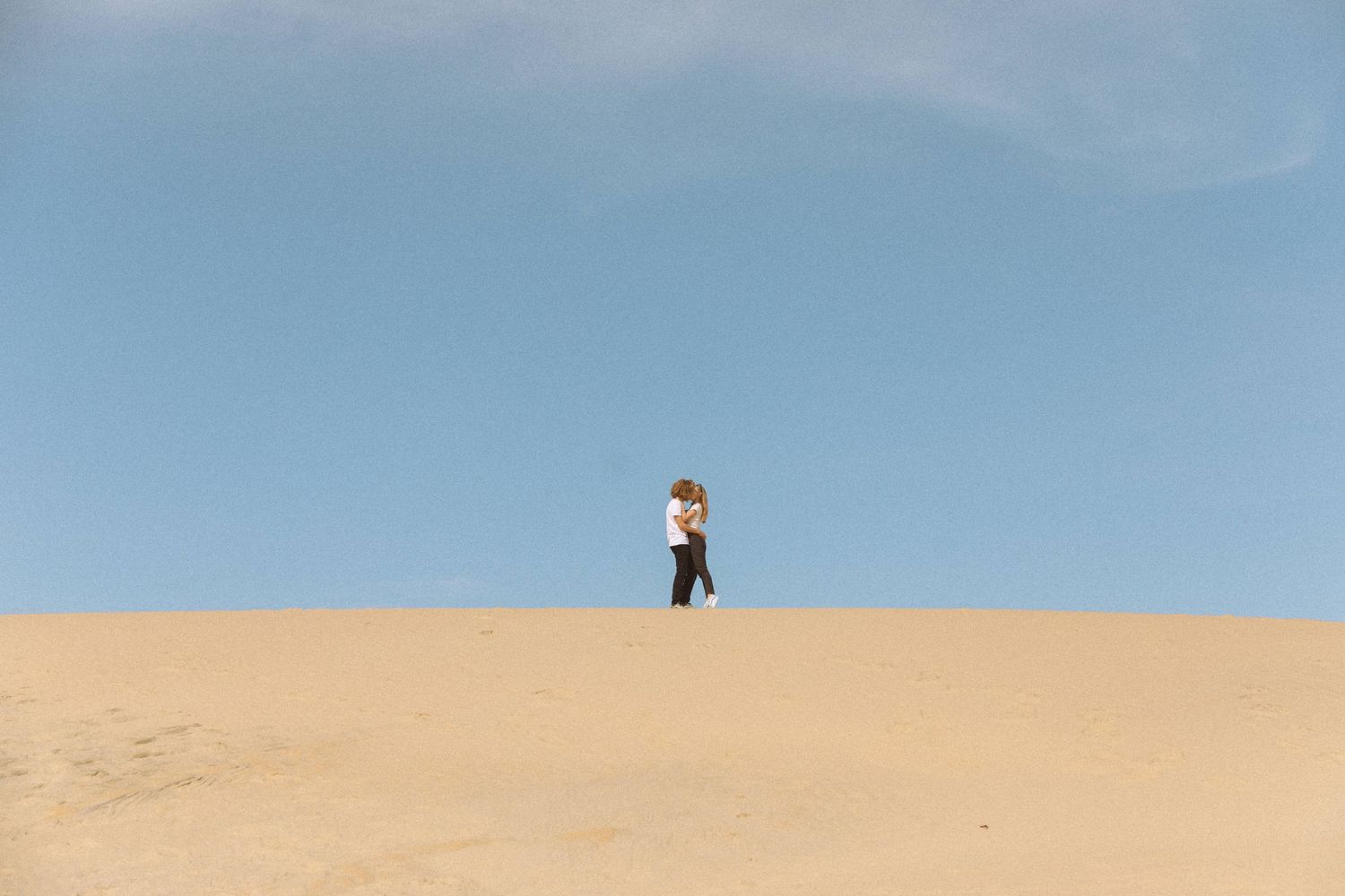 Couple embraces on vast sandy desert dune under clear blue sky in romantic wide-angle engagement photo.