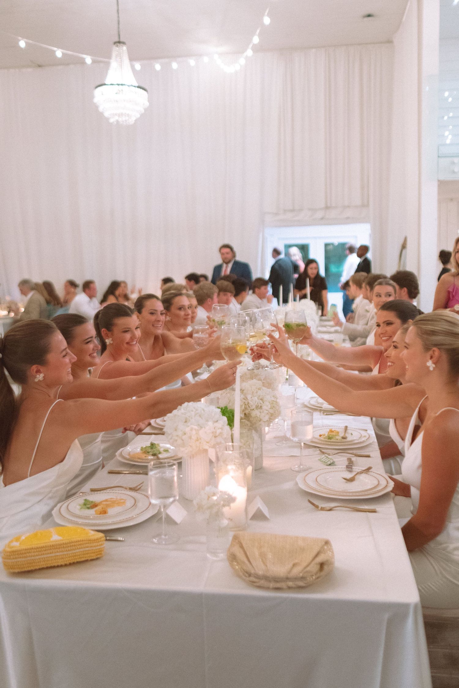Bridesmaids in white dresses raising champagne glasses in elegant toast at wedding reception dinner.