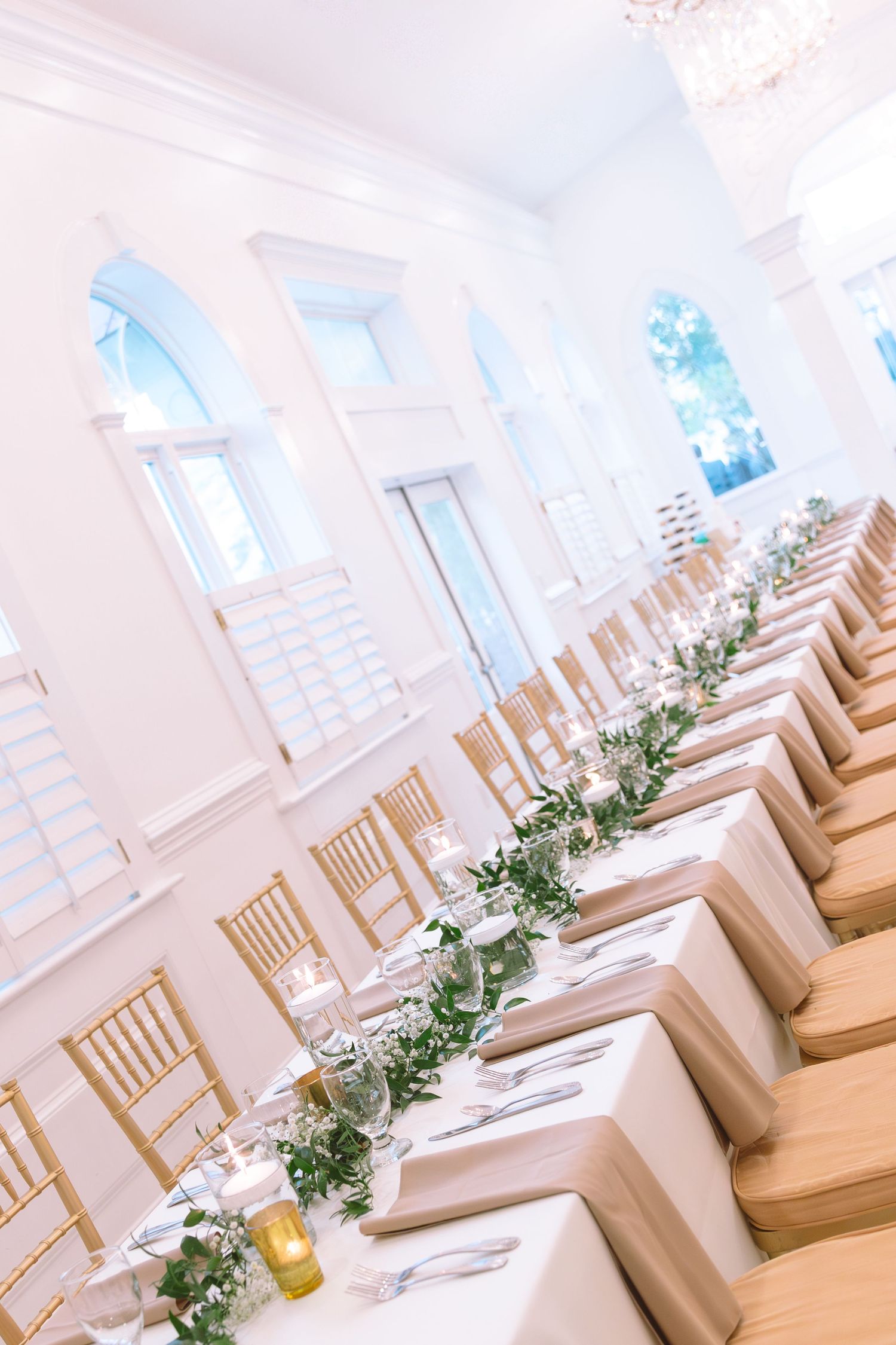 Elegant white wedding reception hall with long tables, gold chairs, and greenery garlands.
