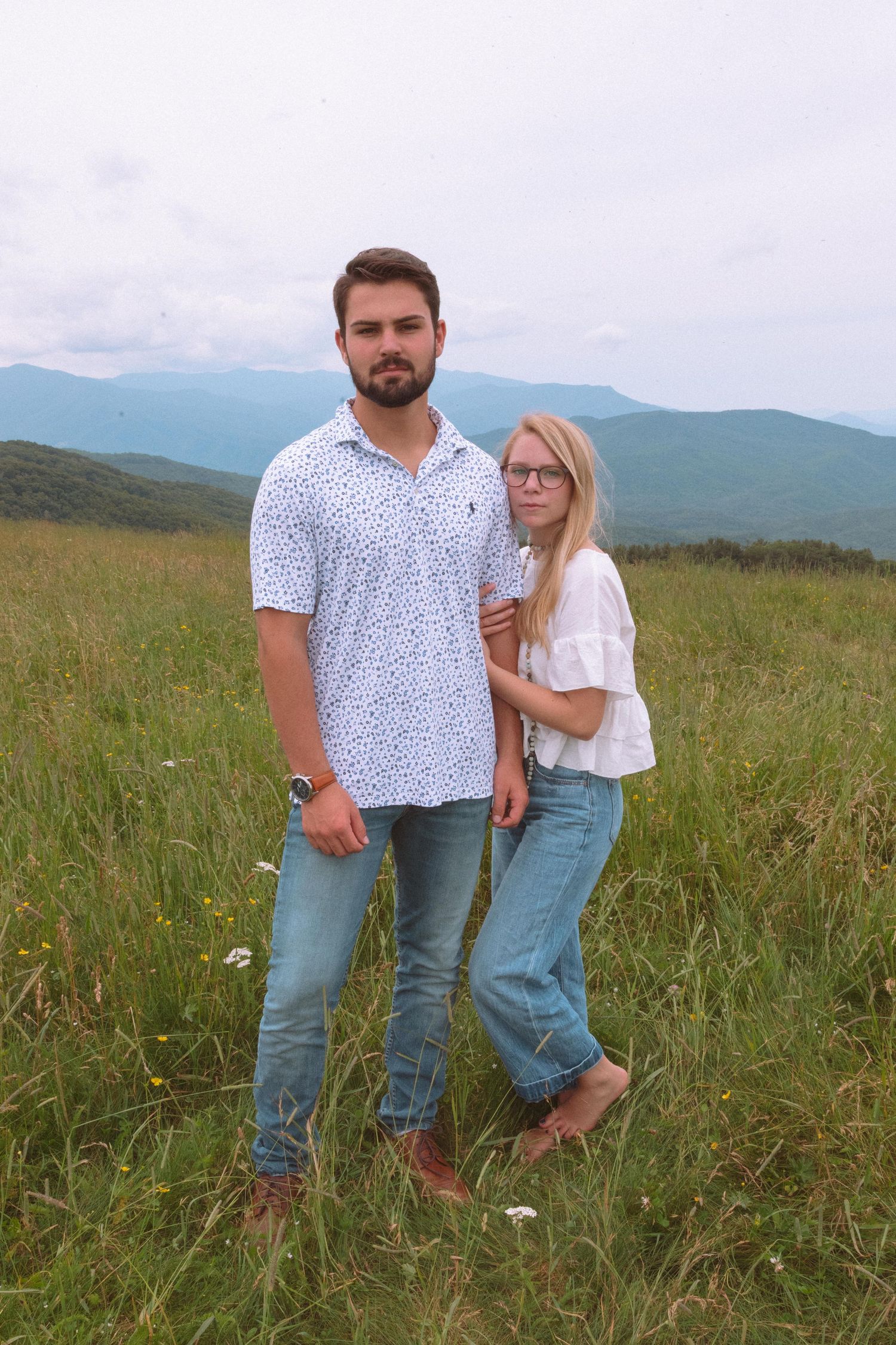 Young couple posing in a green mountain meadow, man in patterned shirt with woman in white top and jeans.