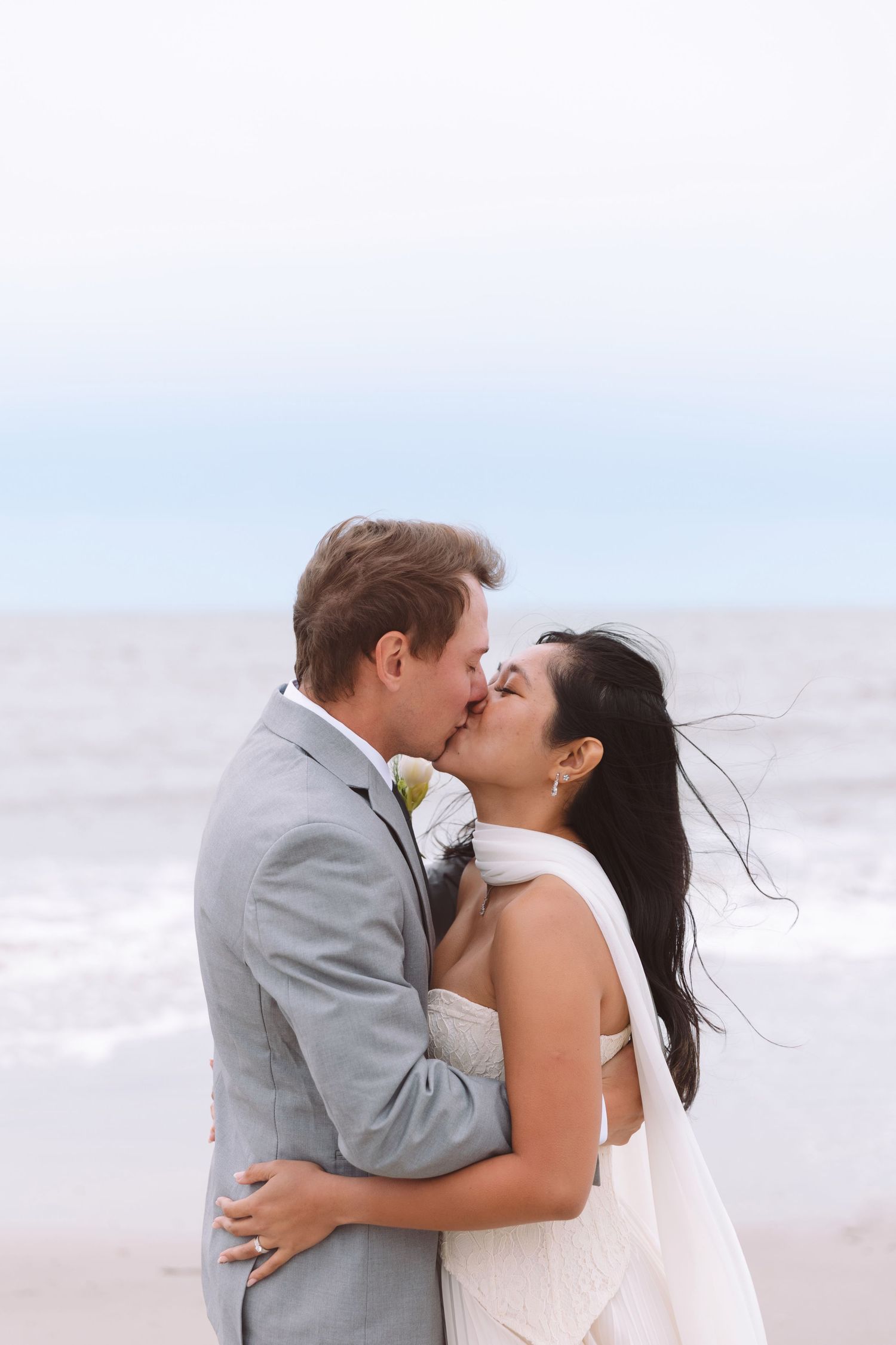 A bride and groom share a romantic kiss on a windy beach, with ocean waves in the background.