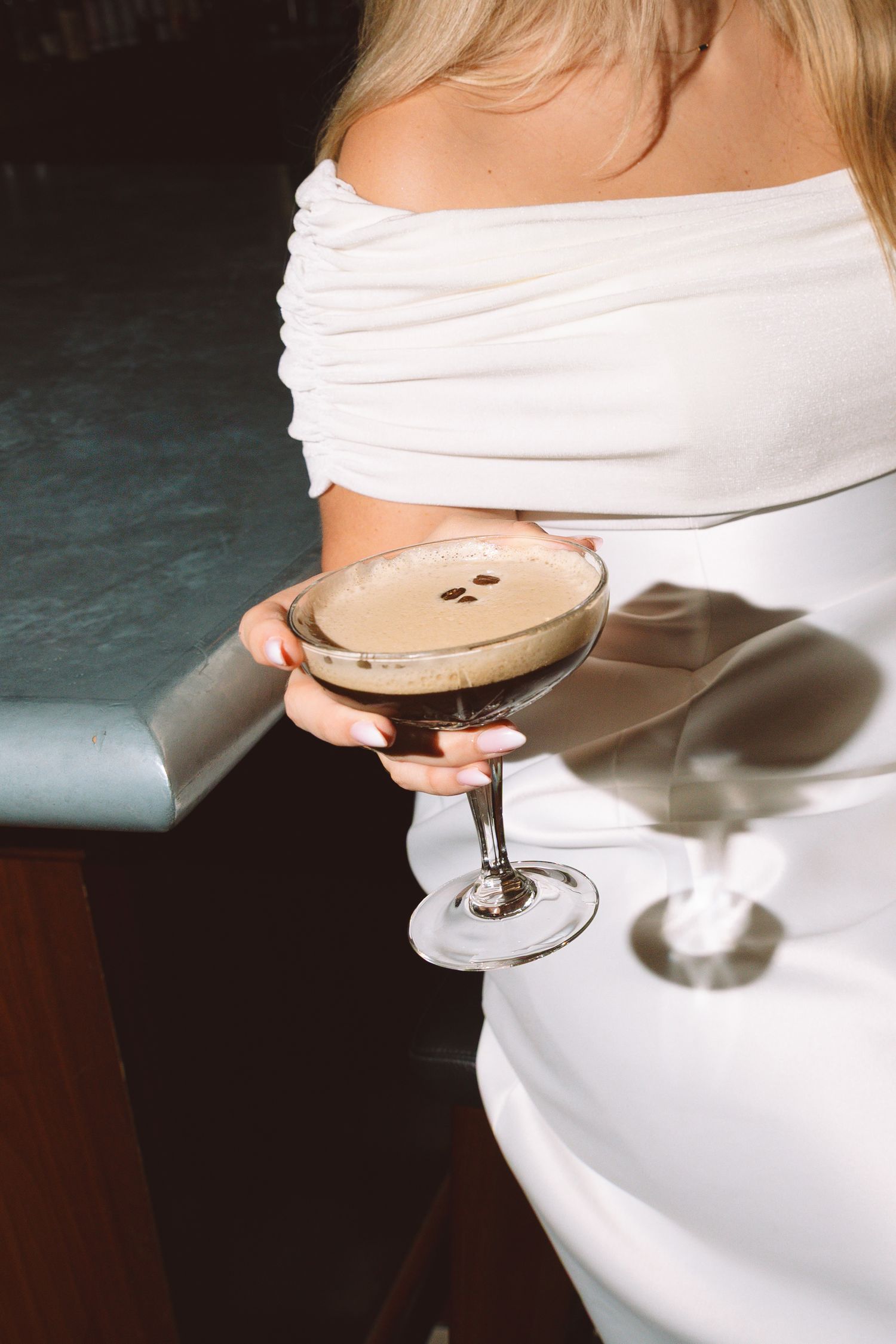 Bride in white off-shoulder dress holds espresso martini cocktail at elegant bar.