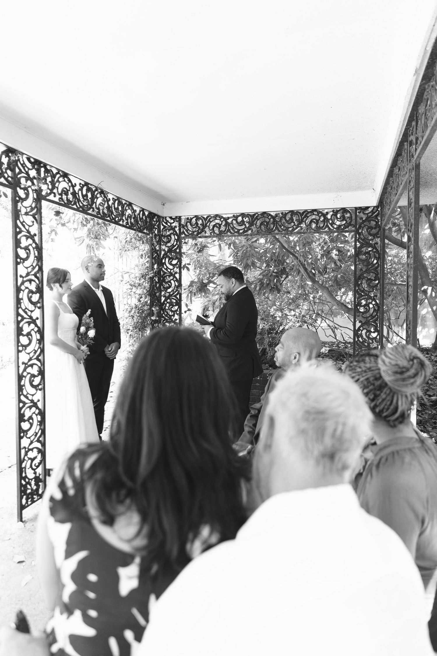 Black and white photo of wedding ceremony with guests watching couple exchange vows under ornate ironwork.