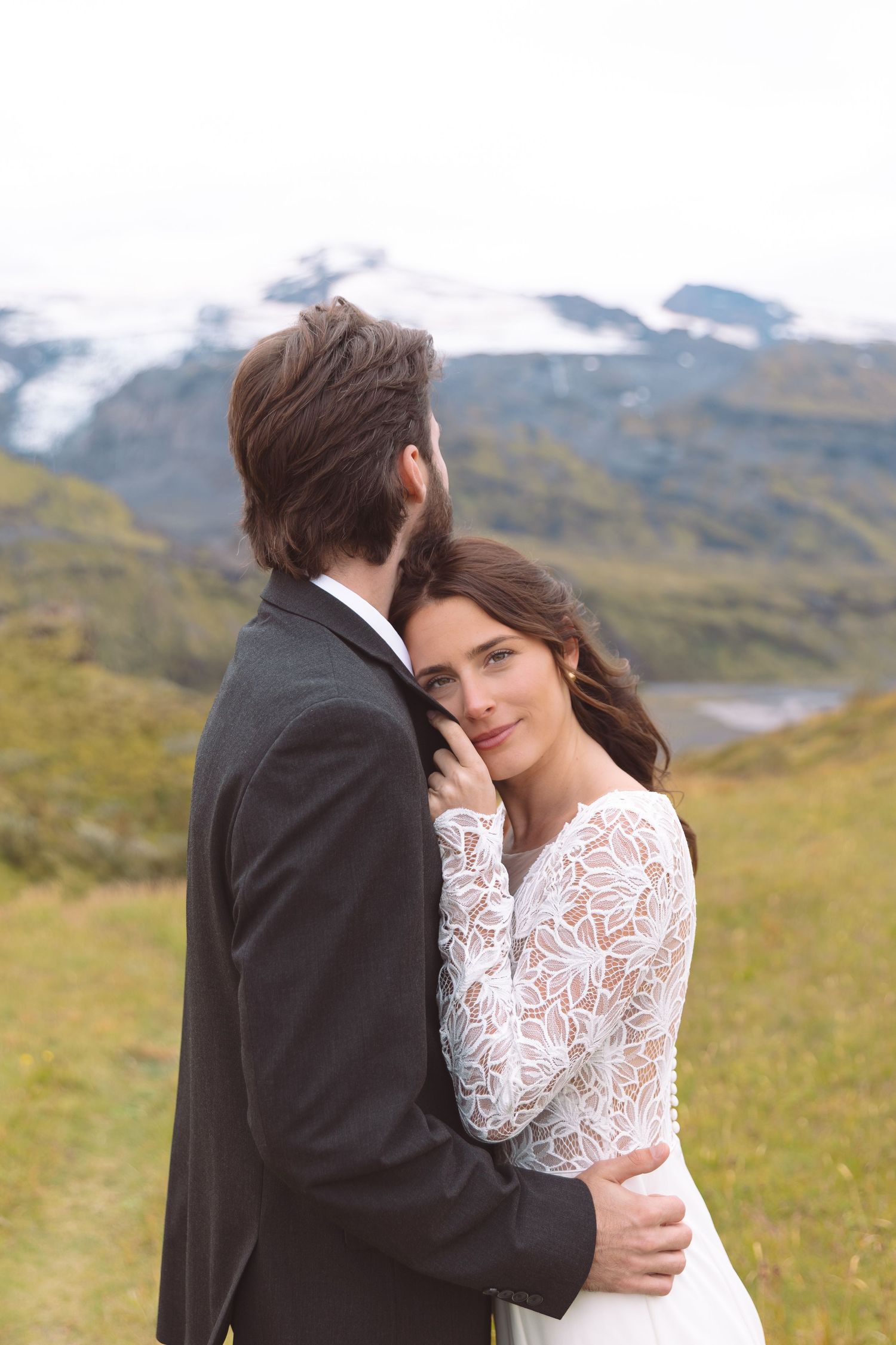 Bride in lace wedding dress resting head on groom's chest smiling softly with Icelandic mountains backdrop.