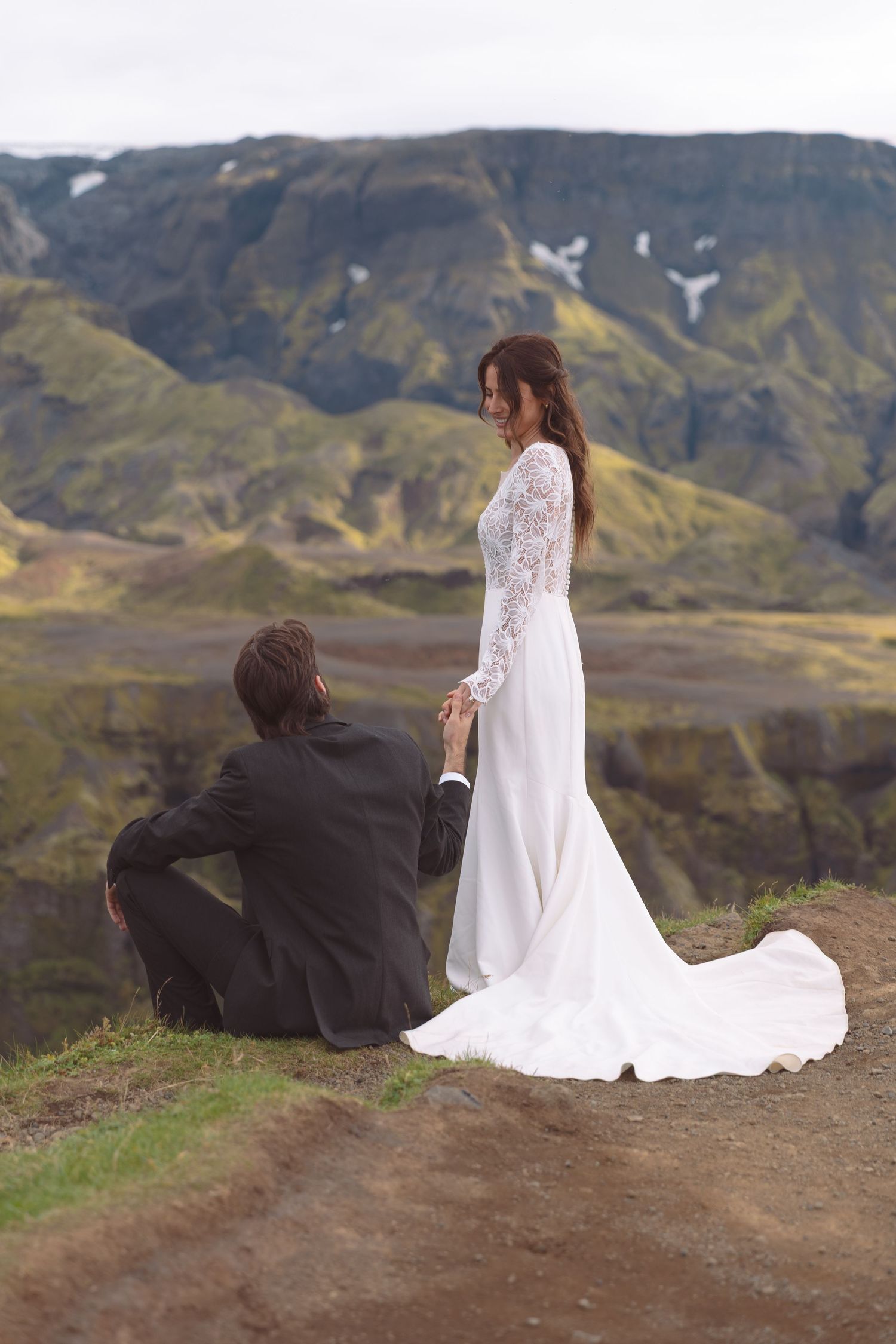 Groom sits on Iceland hillside holding bride's hand as she stands in elegant lace wedding gown.