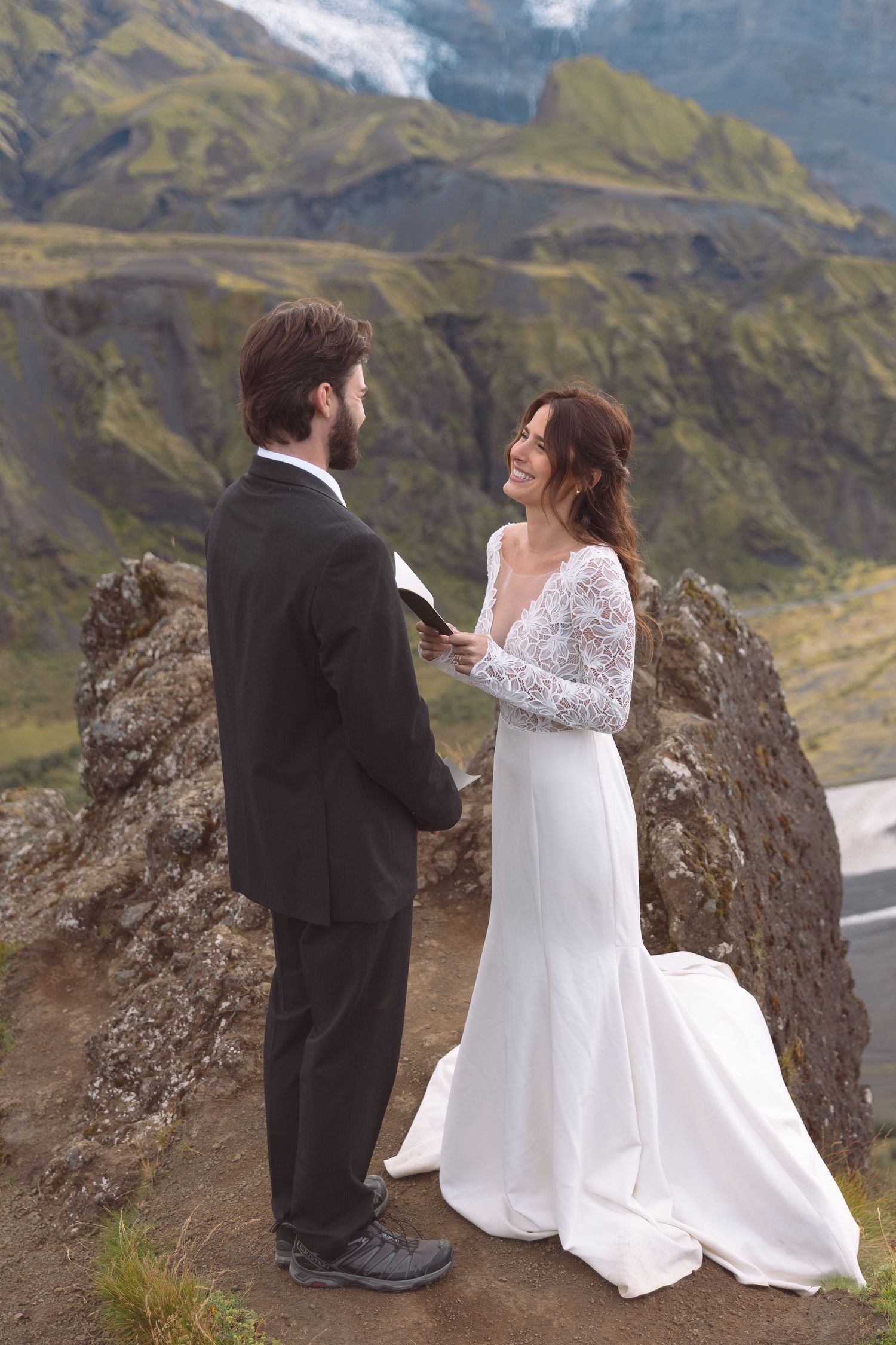 Groom reads vows to smiling bride in lace dress during intimate mountain elopement ceremony in Iceland.