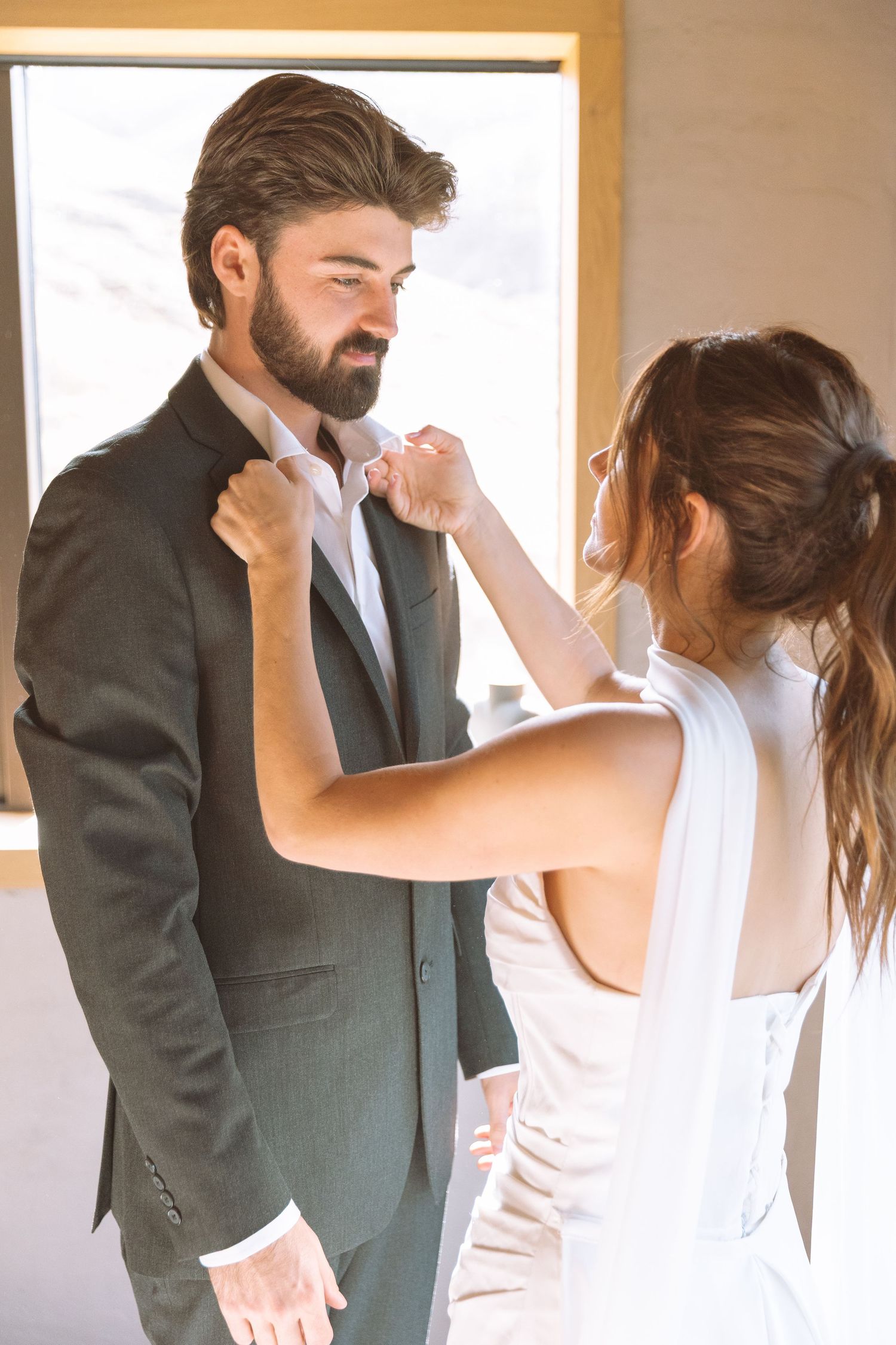 Bride helping groom with his suit jacket near a bright window on wedding day.