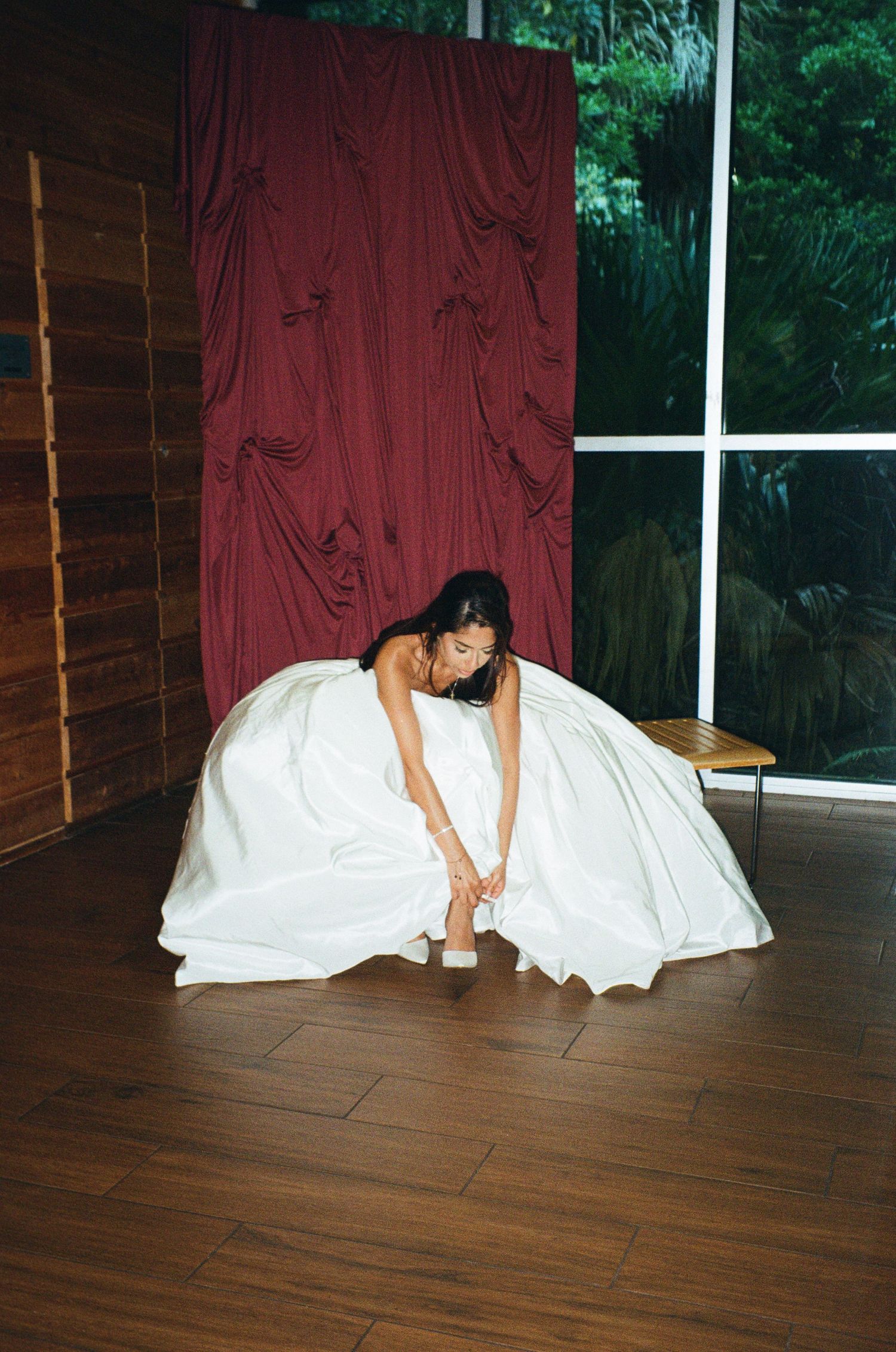 Bride in a white ball gown adjusting her shoe on a wooden floor, with a red curtain backdrop.