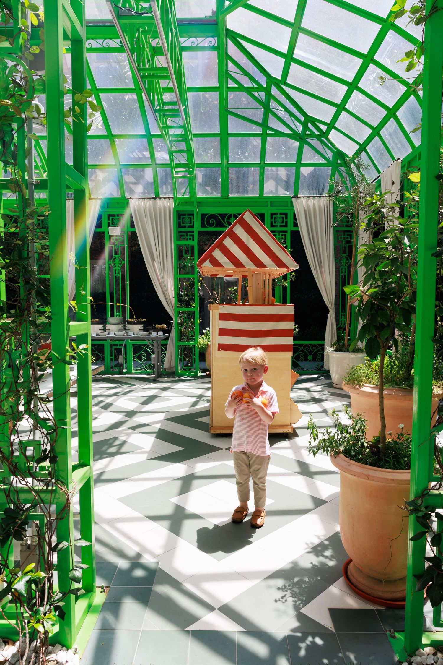 A young blonde child stands in a bright green greenhouse corridor holding food near a striped red and white kiosk.