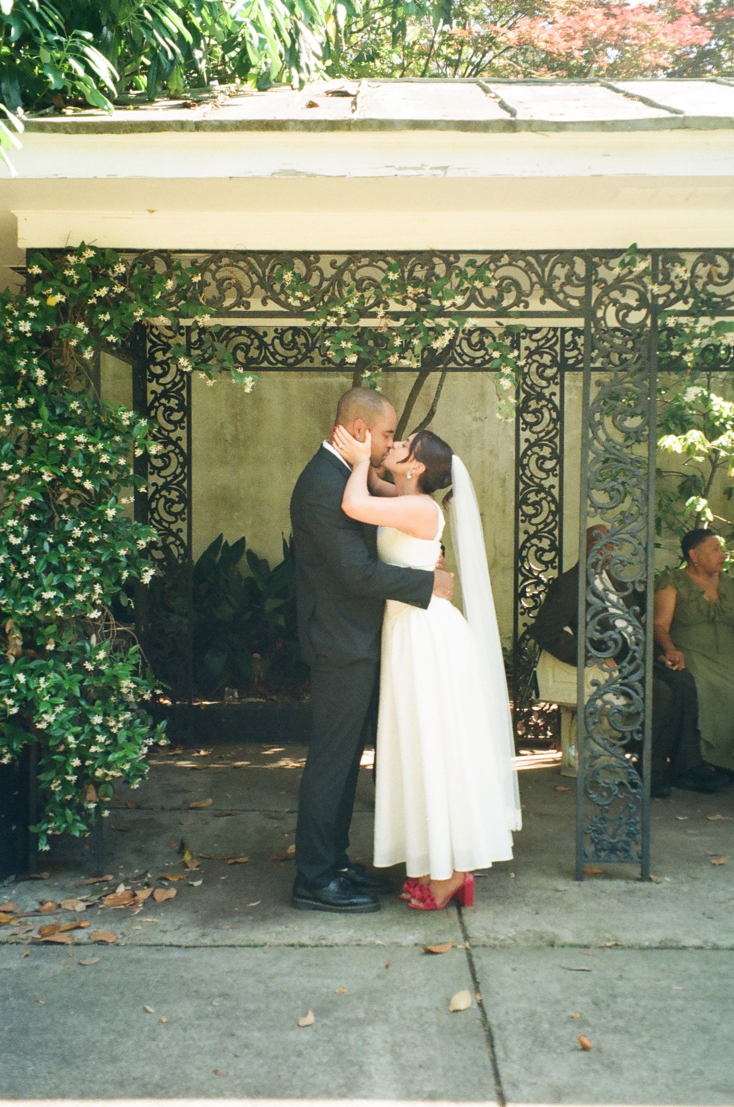 Bride and groom share first kiss under ornate iron gate surrounded by lush greenery at outdoor wedding ceremony.