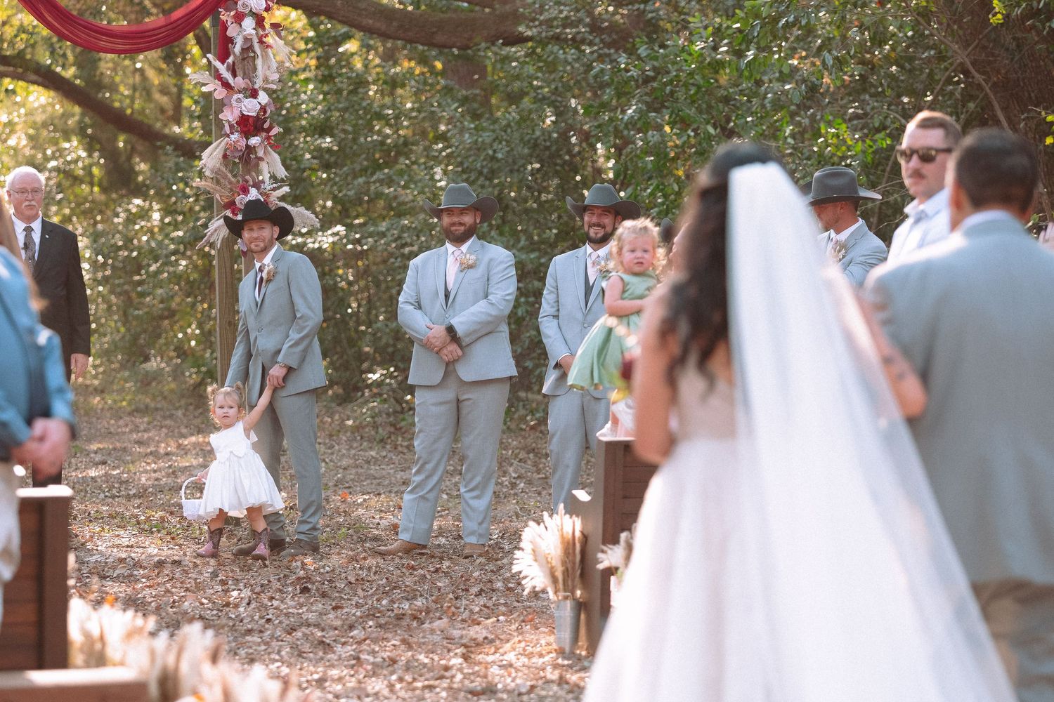 Groom and groomsmen in gray suits await bride at outdoor rustic wedding ceremony aisle.