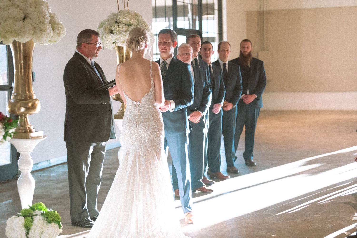 Bride in backless lace gown stands at altar with groomsmen in grey suits during wedding ceremony.