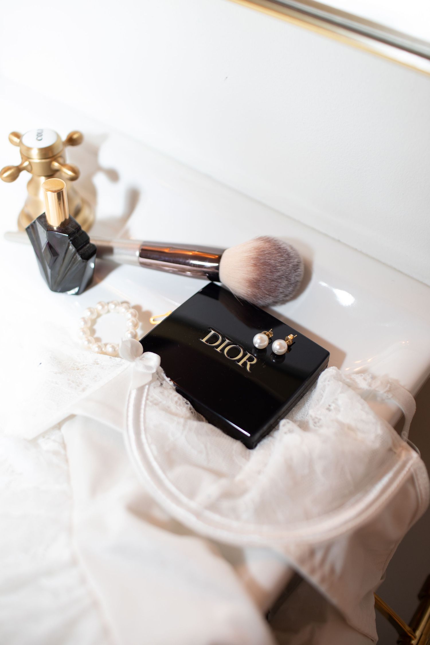 Dior makeup compact with pearl earrings, brush, and perfume bottle on white lace wedding dress.