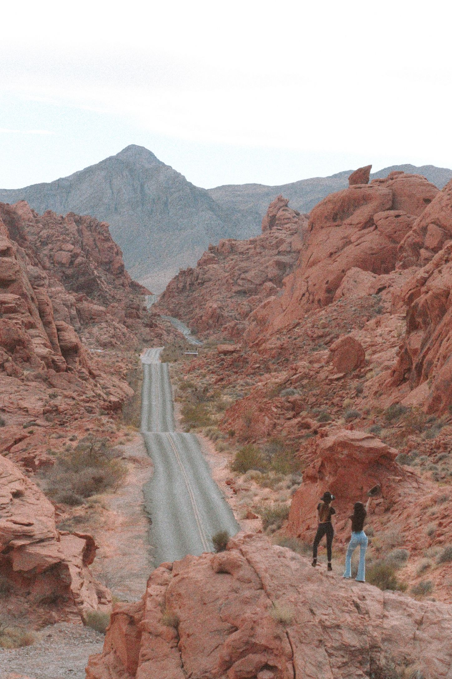 Two people standing on red rock formations overlooking a desert road.