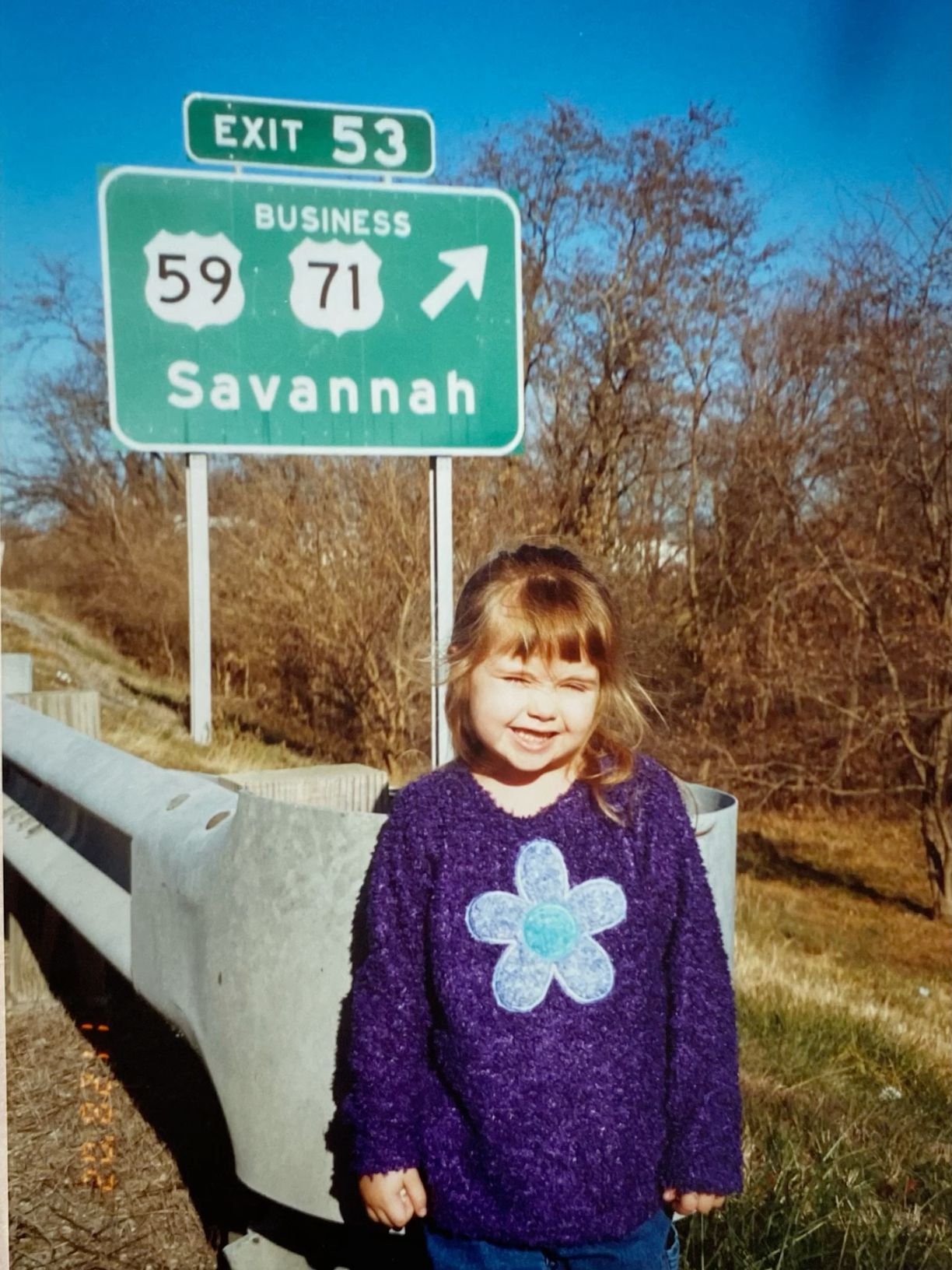 A young girl in a purple floral sweater smiling in front of an Exit 53 Savannah highway sign.