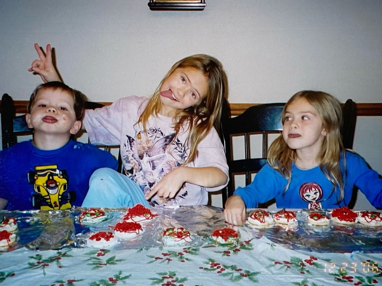 Three children smile at a table decorated with festive Christmas cupcakes and holiday tablecloth.