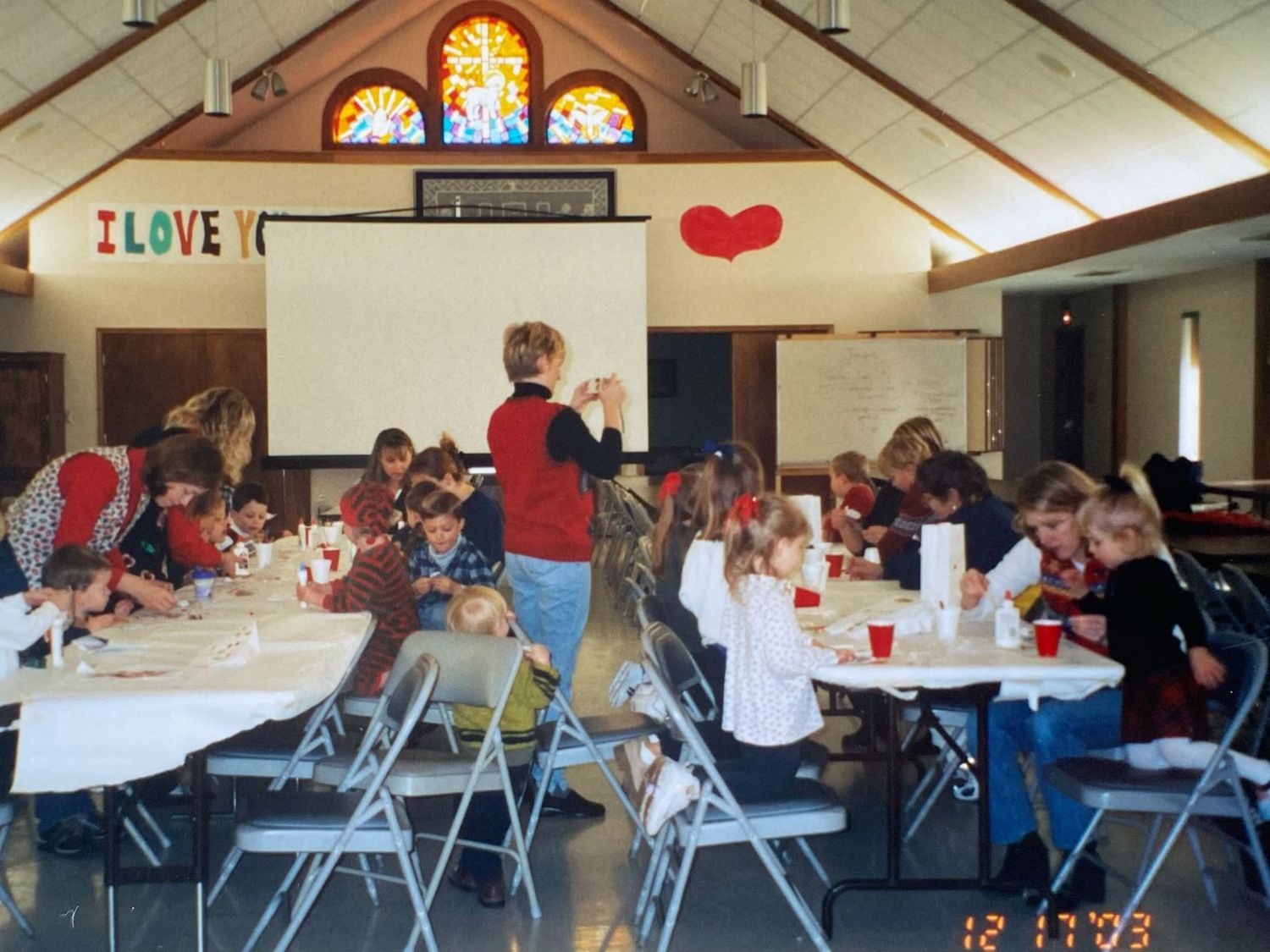 Children and adults crafting at tables in a decorated church hall with I Love You banner.