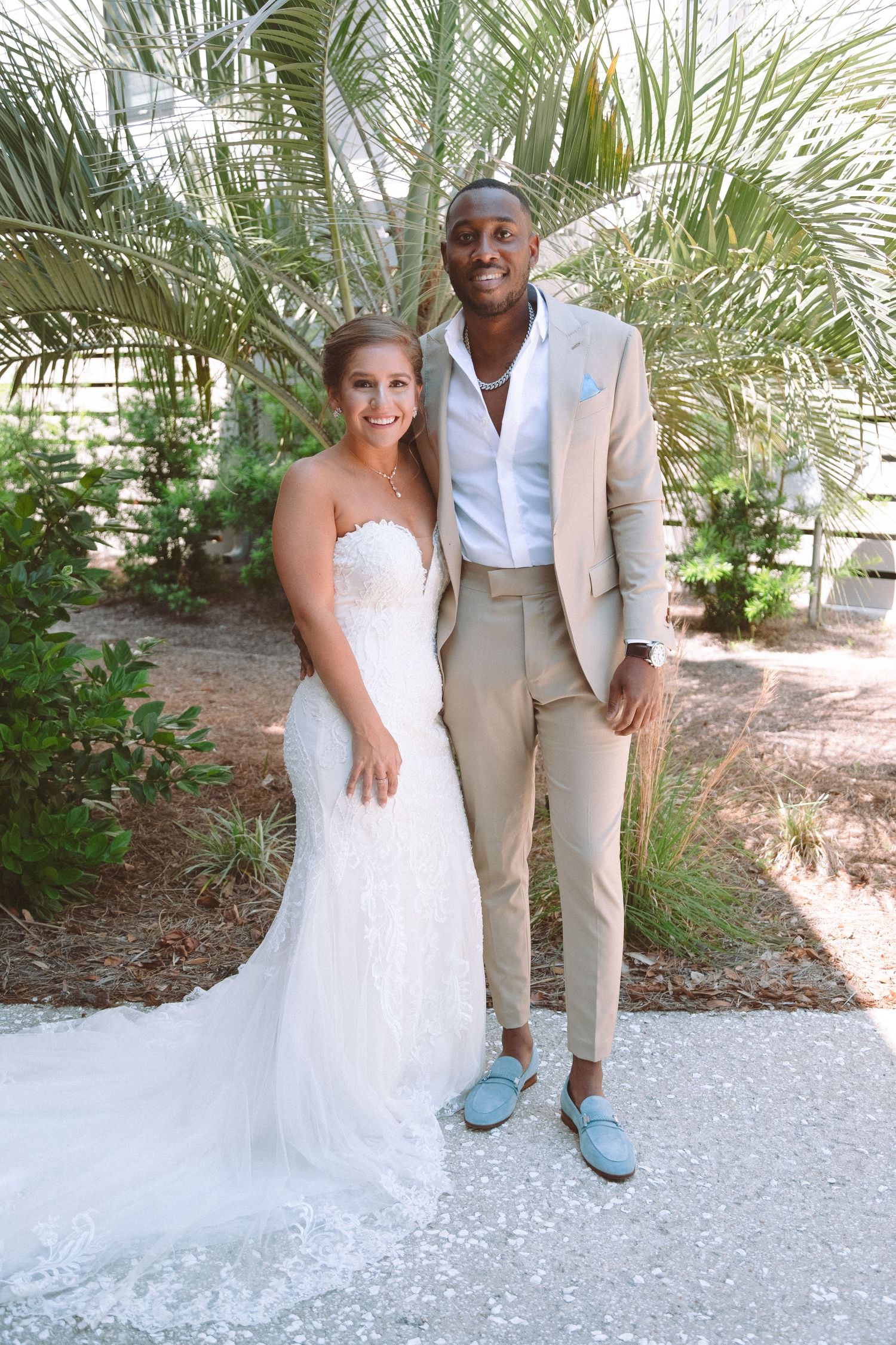Bride and groom smiling together outdoors, she in white gown, he in tan suit with blue loafers.