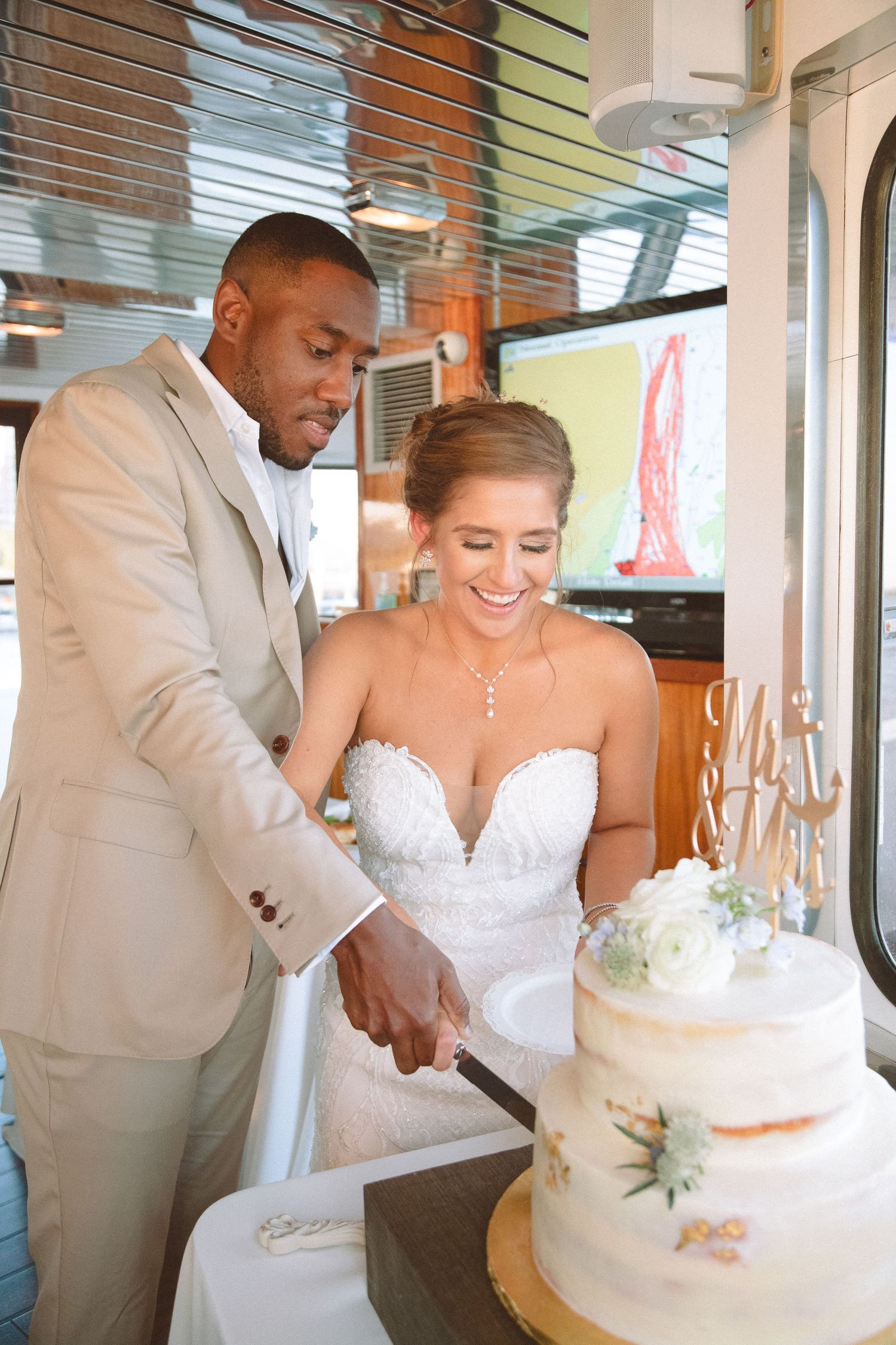 Smiling bride and groom in tan suit cutting elegant two-tier wedding cake at boat reception.