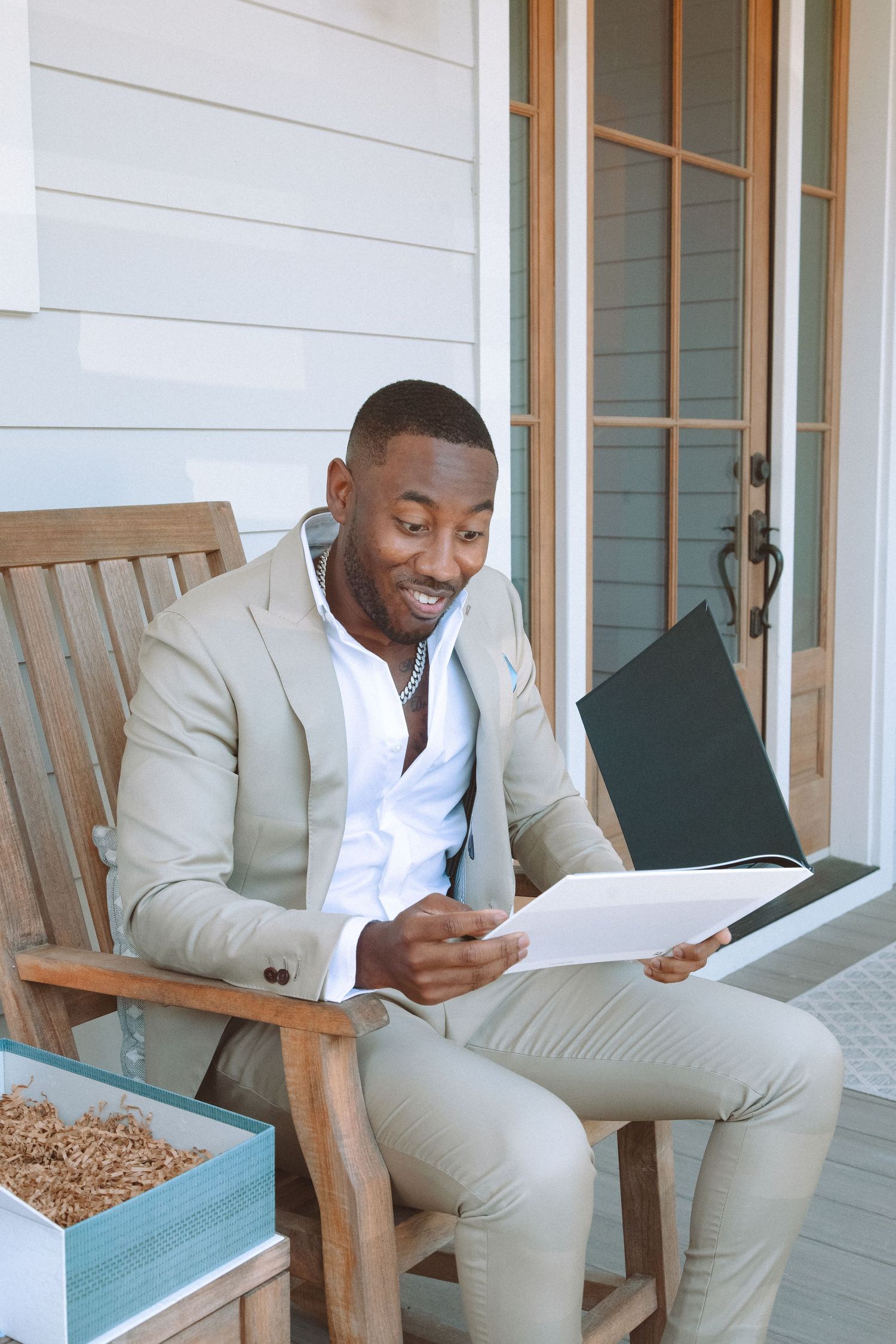 Smiling man in beige suit happily reading a black photo album while seated on outdoor wooden chair.
