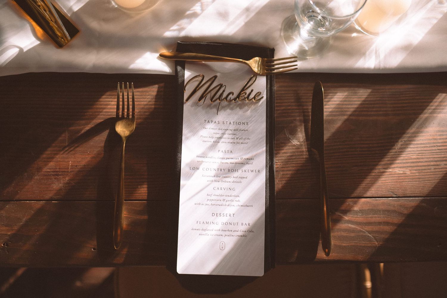 Personalized wedding menu card with gold script name on white linen with gold flatware on wooden table.