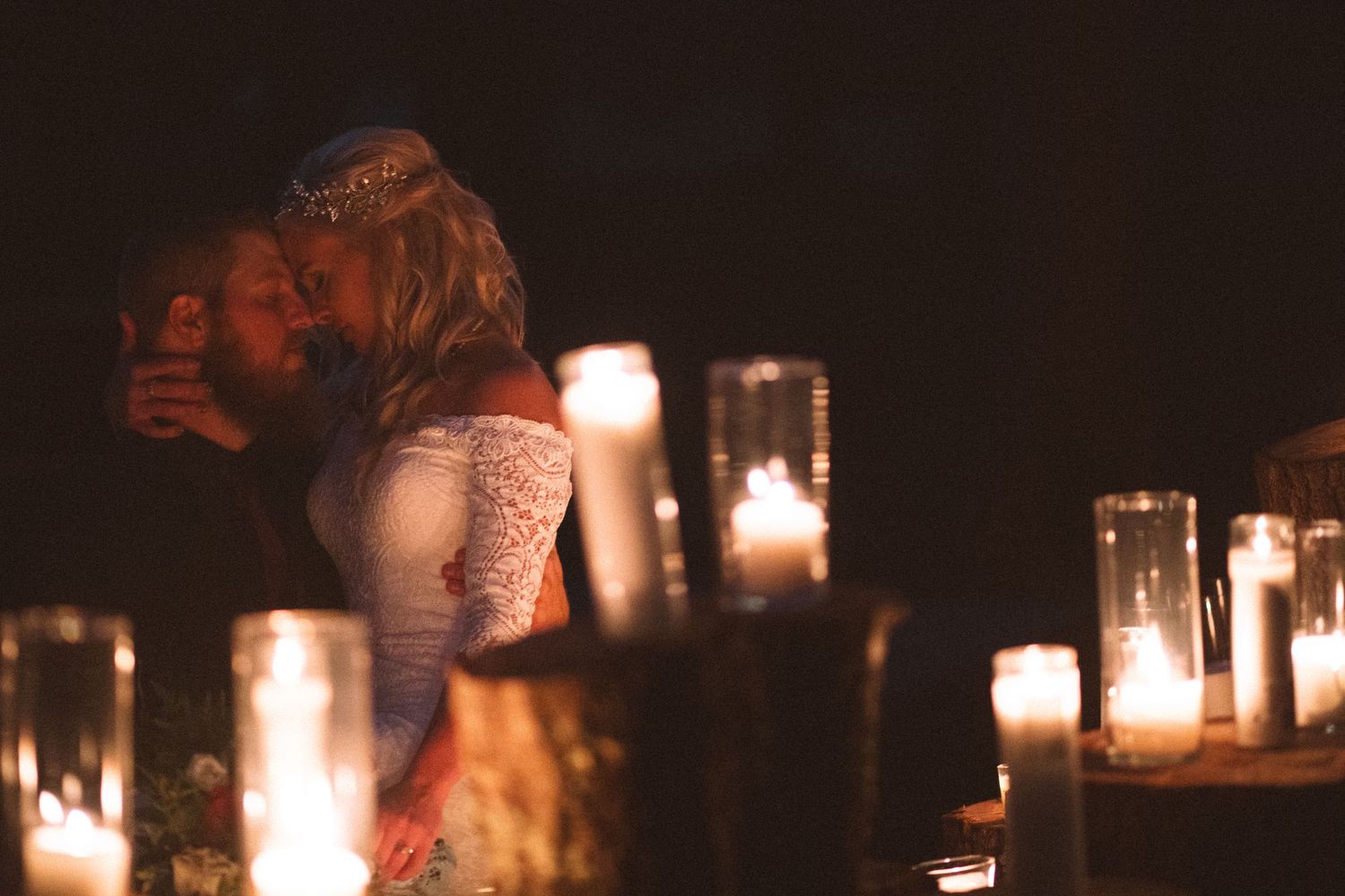 Bride and groom share a romantic candlelit kiss surrounded by glowing candles in a dark, intimate wedding setting.