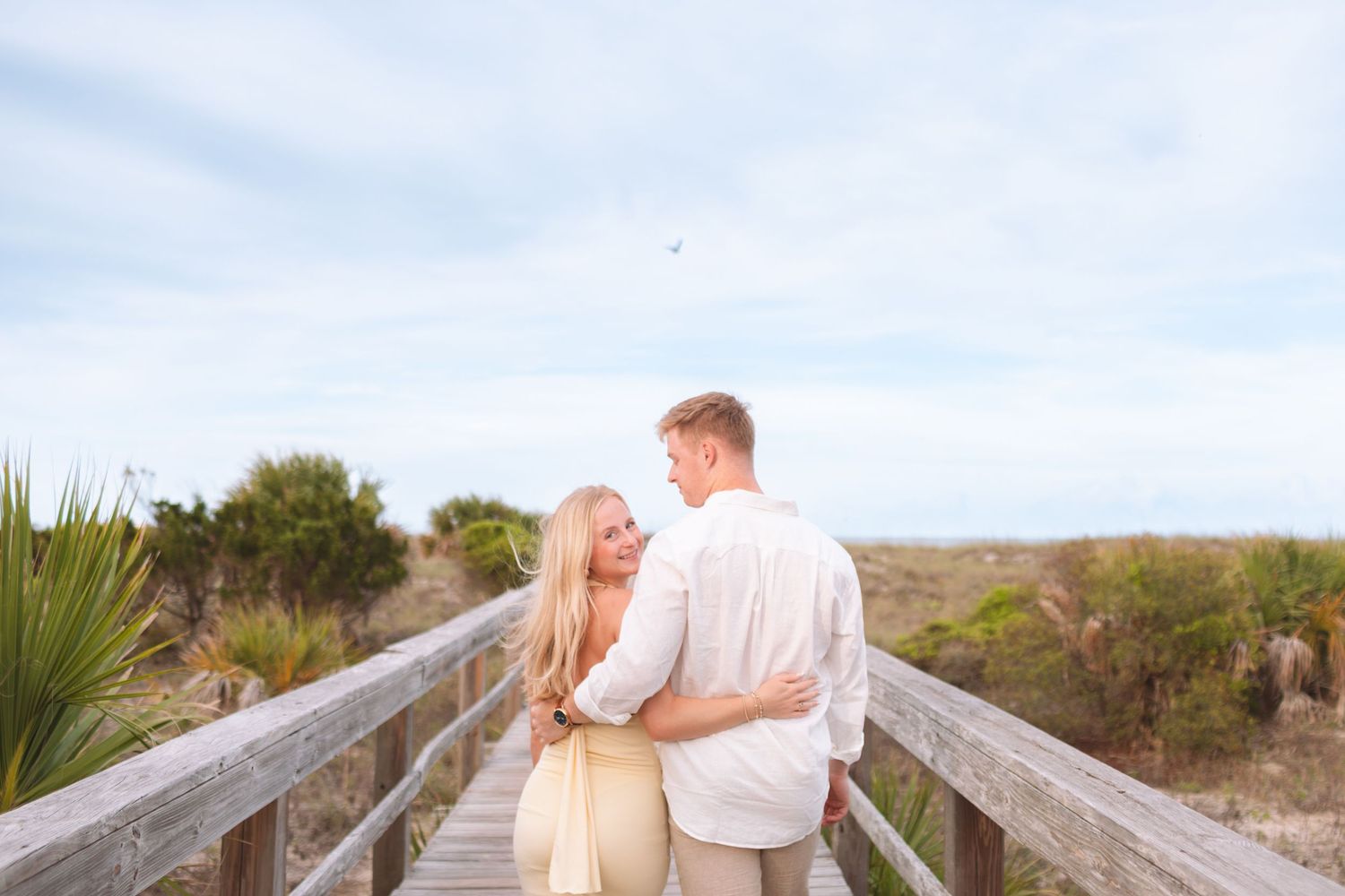 Romantic couple embracing on a wooden boardwalk overlooking sandy beach dunes under a cloudy sky.