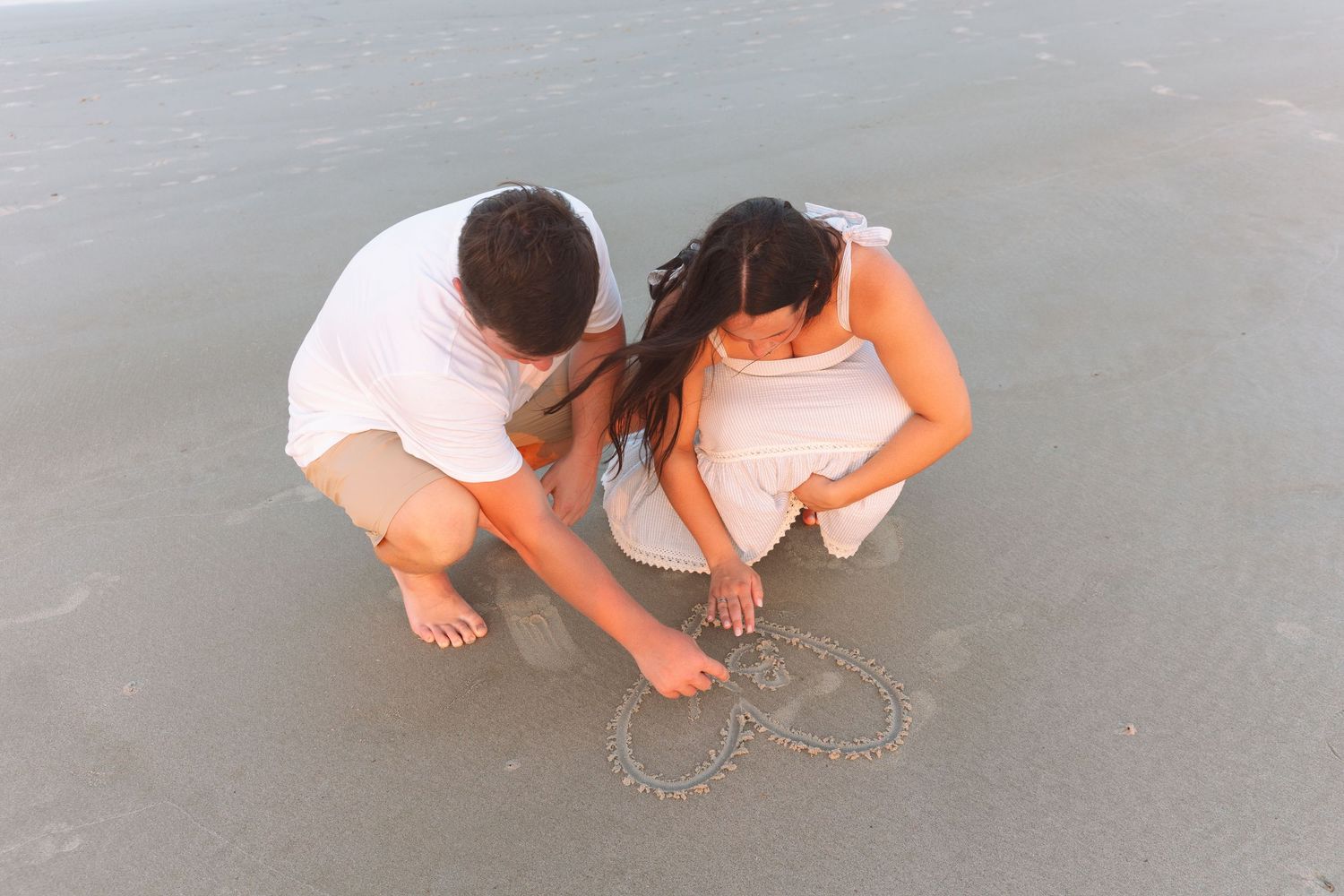 Couple crouching on wet sand at the beach, drawing a heart shape together near the ocean shoreline.