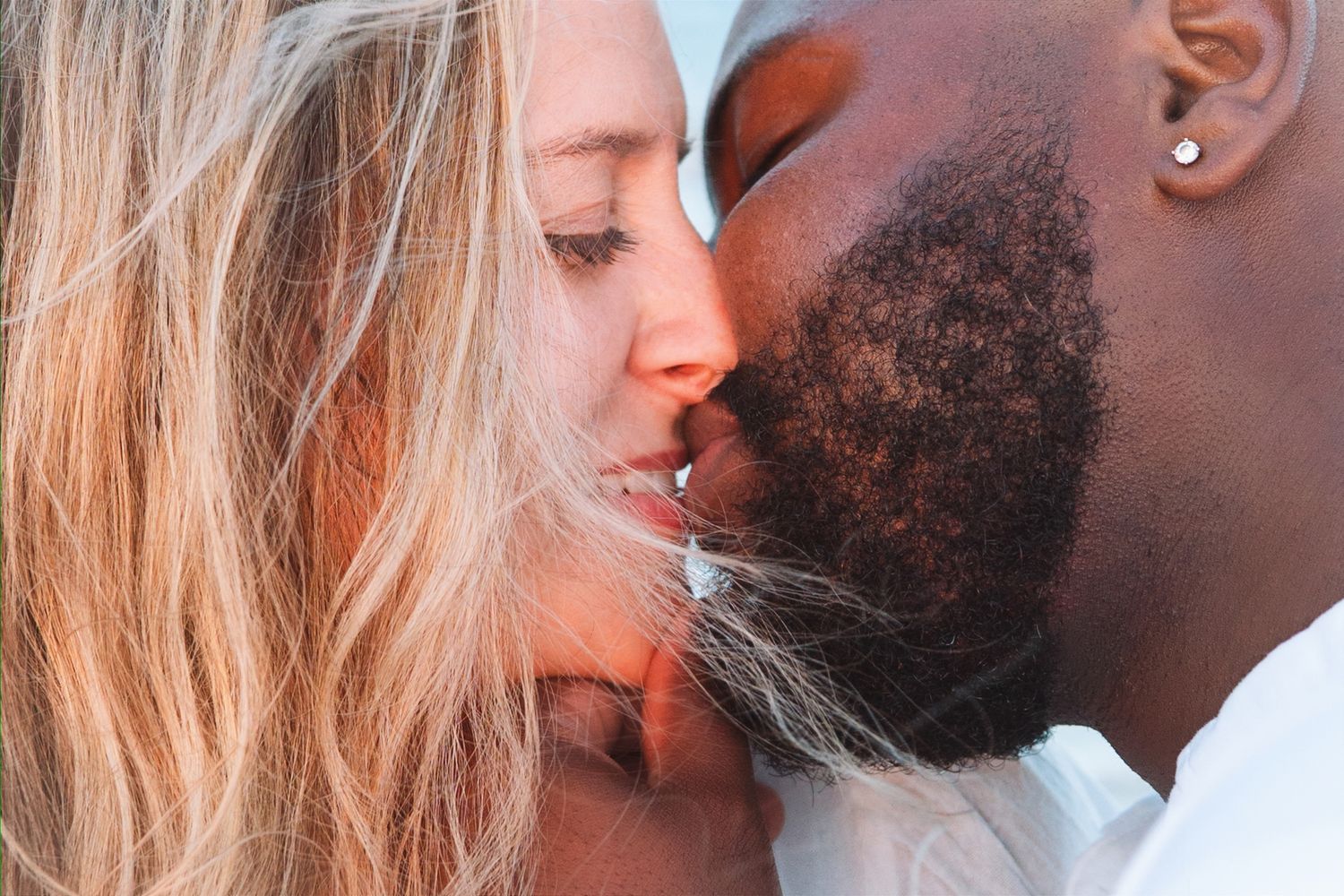 Interracial couple sharing a romantic kiss in a close-up outdoor photo.