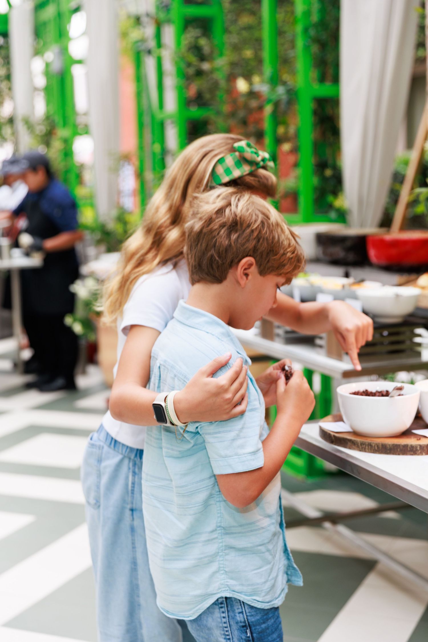 A girl with a green bow hugs a boy as they explore a food station at an outdoor event with green decor.