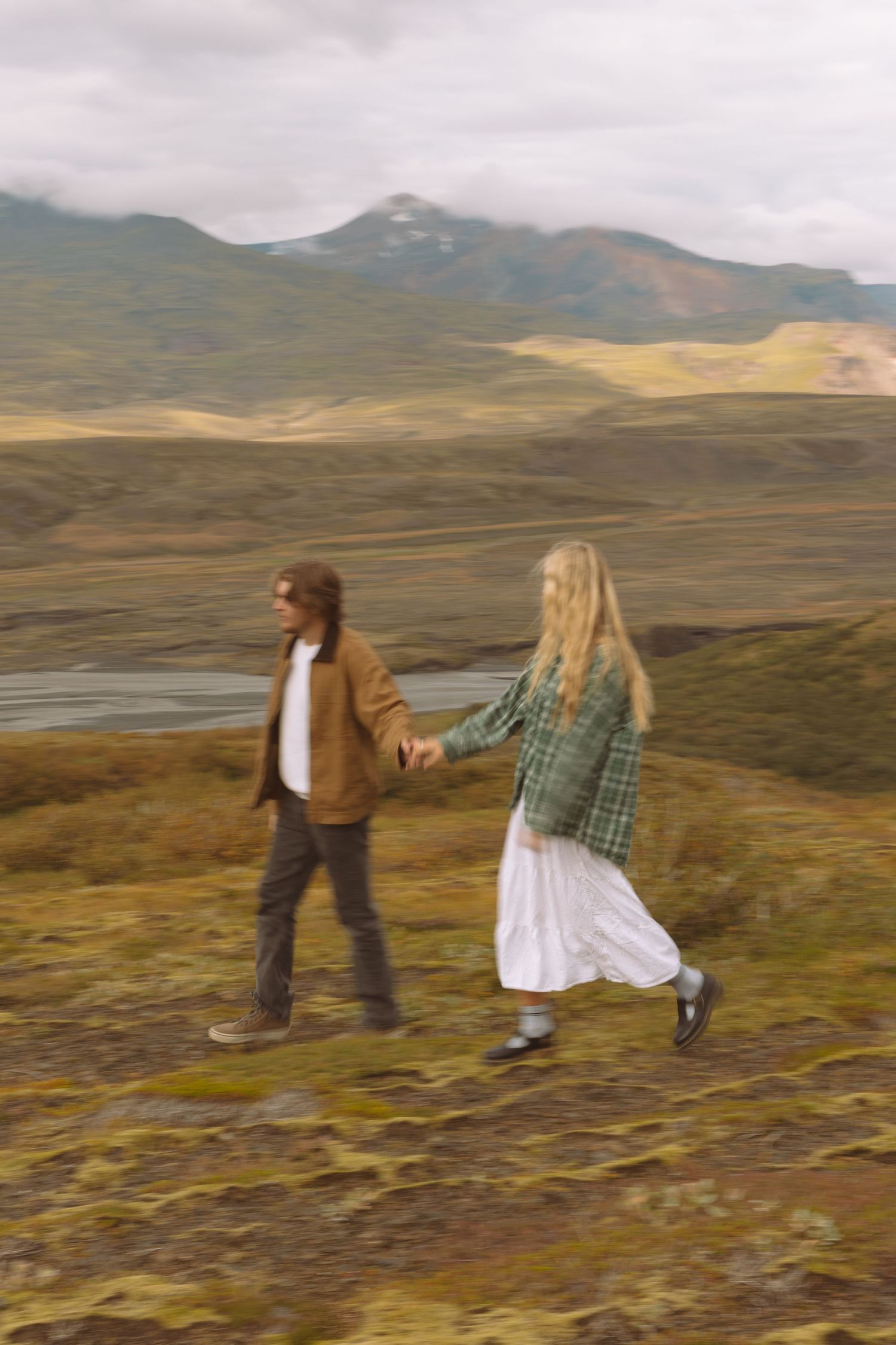 Couple holds hands while walking through a dramatic Icelandic highland landscape with mountains behind.