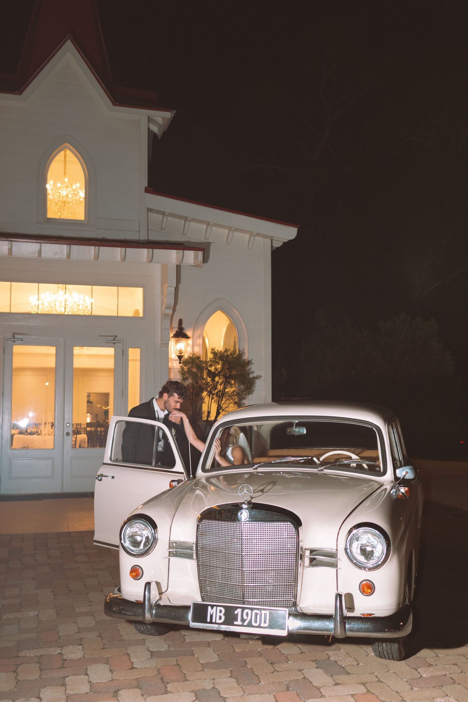 Newlyweds kissing beside vintage Mercedes outside illuminated wedding venue at night.
