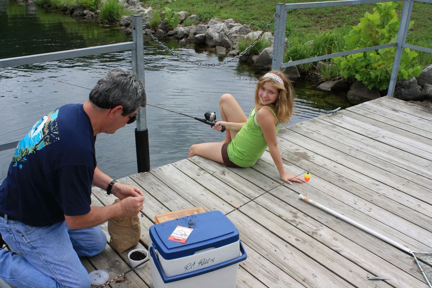 A man and a teenage girl fishing together on a wooden dock beside a calm lake on a sunny day.