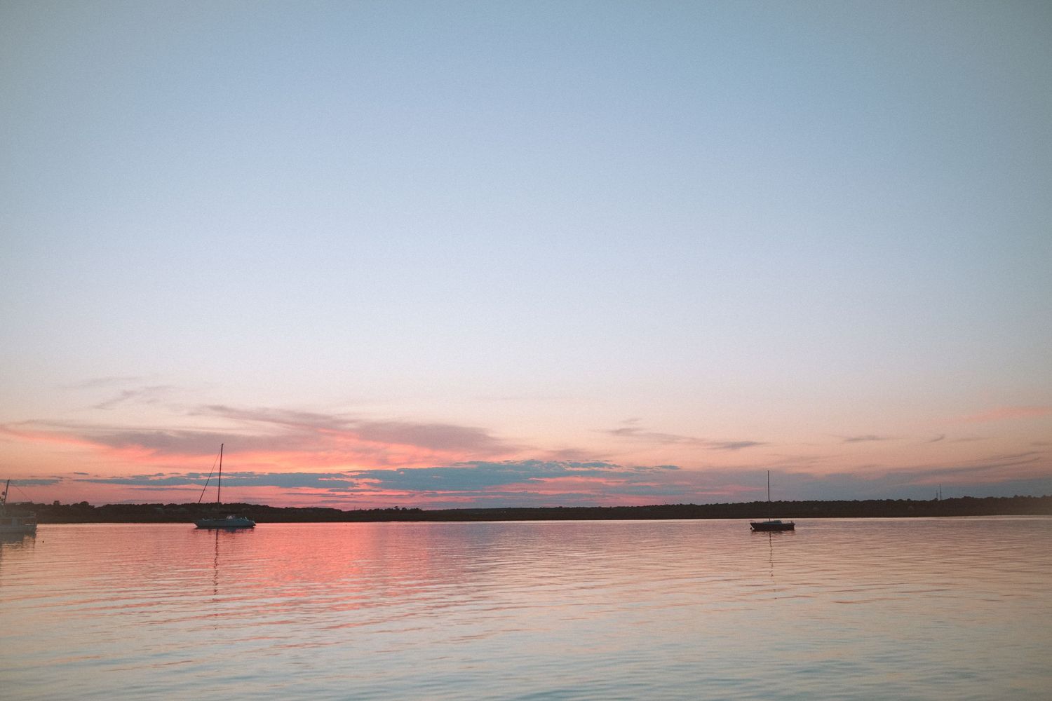 Serene waterfront sunset with soft pink hues reflecting on calm water and distant sailboats.