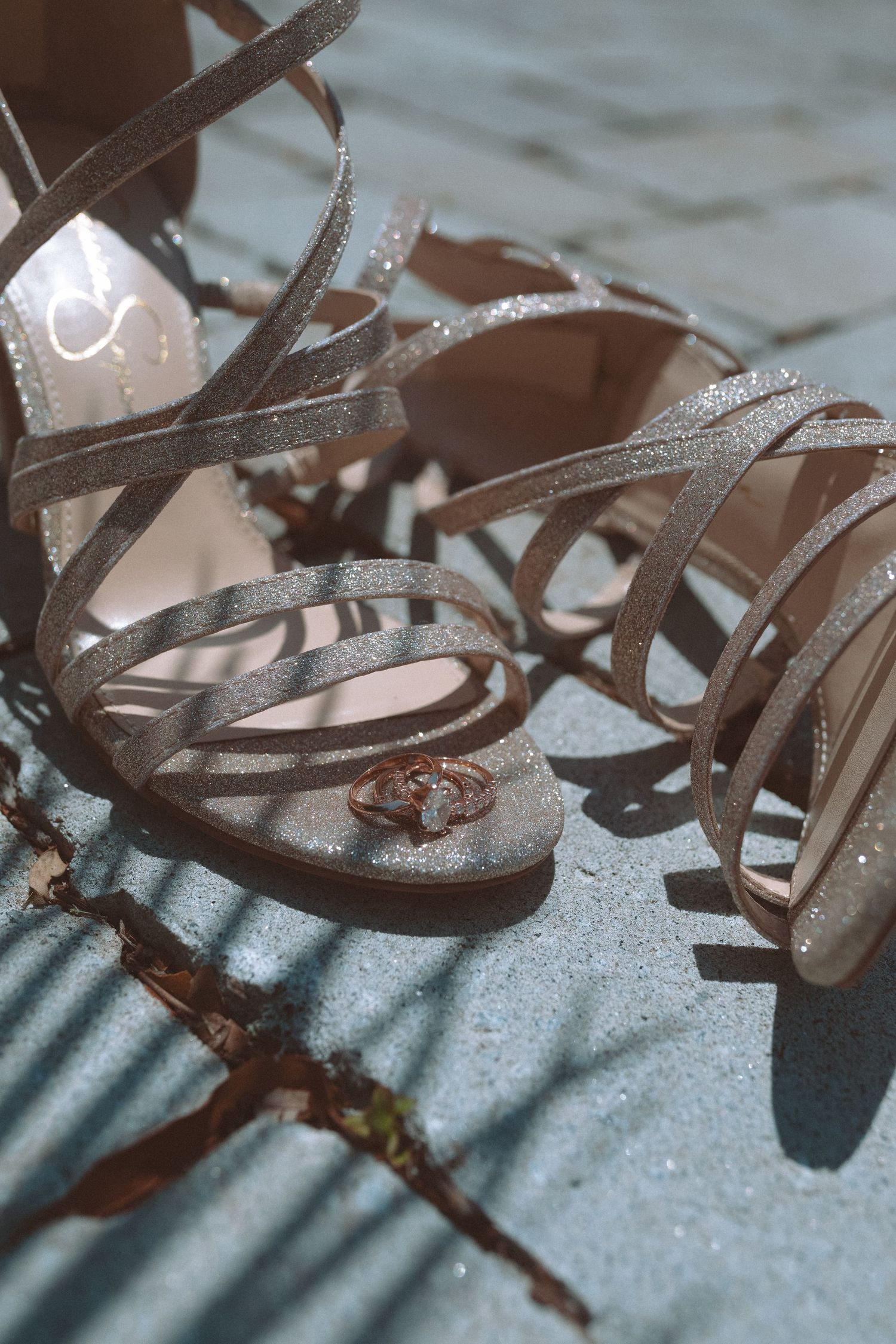 Sparkling strappy silver wedding heels with diamond engagement ring placed between the shoes outdoors.
