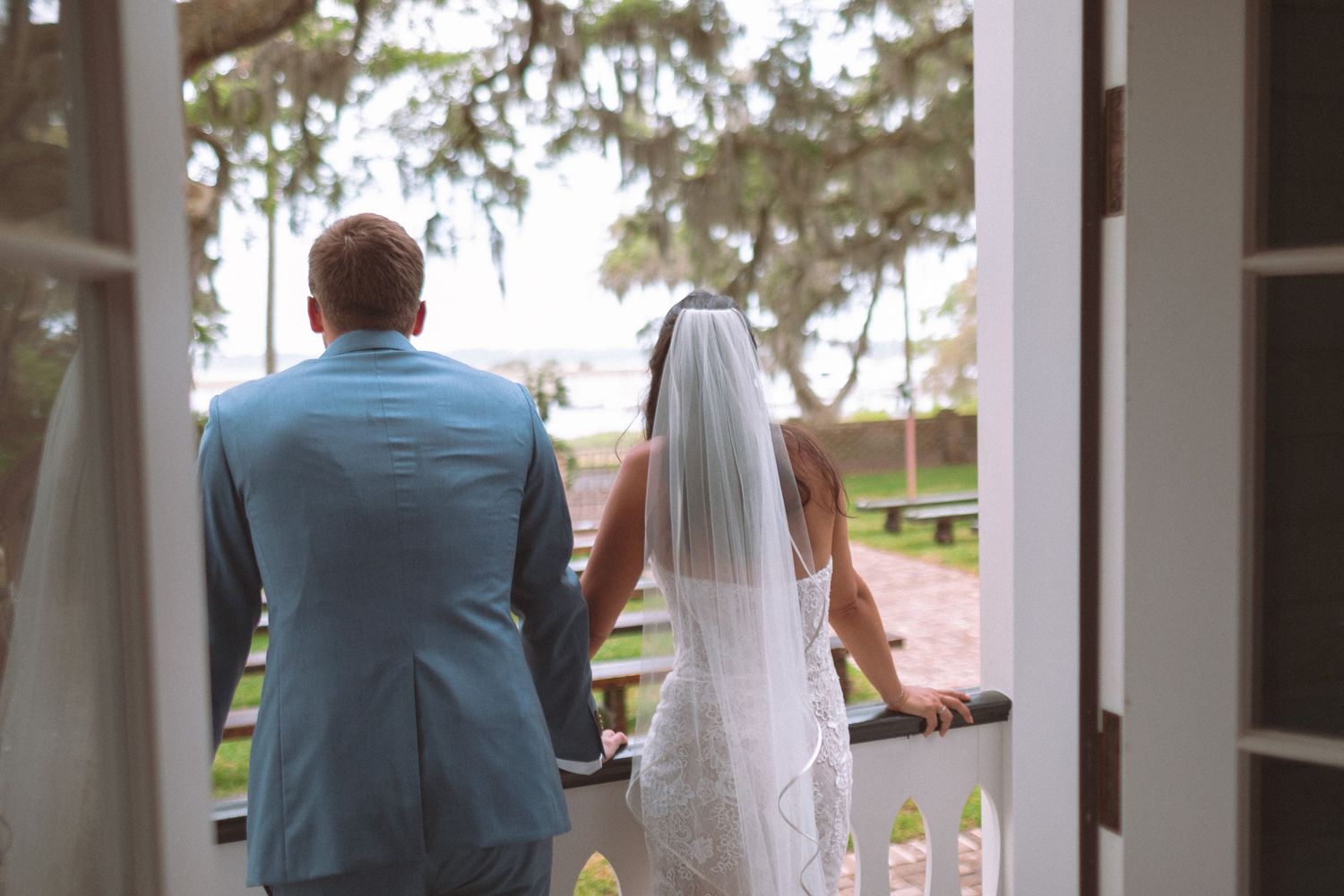 Bride and groom in light blue suit walk hand in hand through white doors toward waterfront.