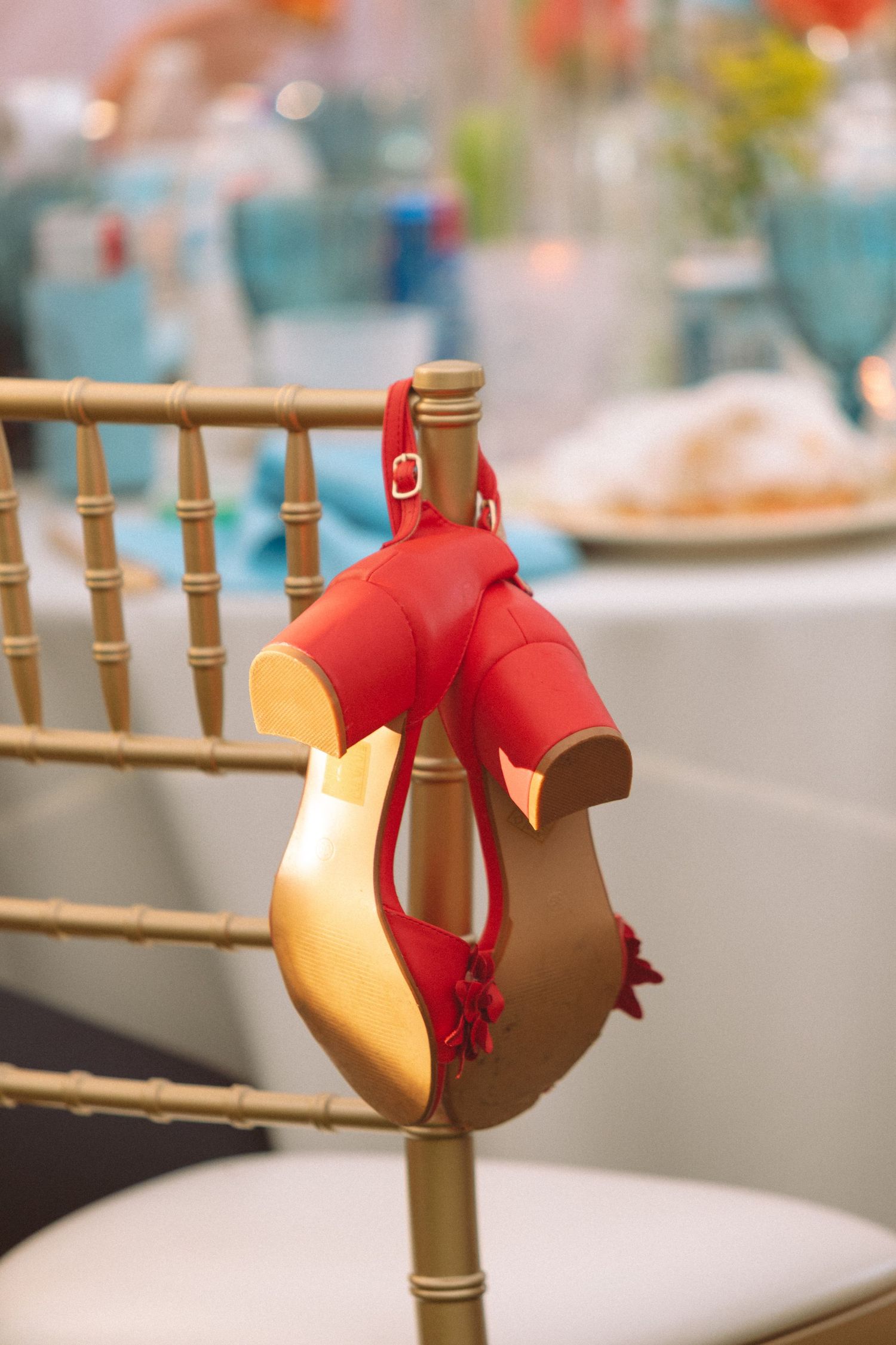Red high heel bridal shoes with ankle straps hanging on a gold chiavari chair at a wedding reception.