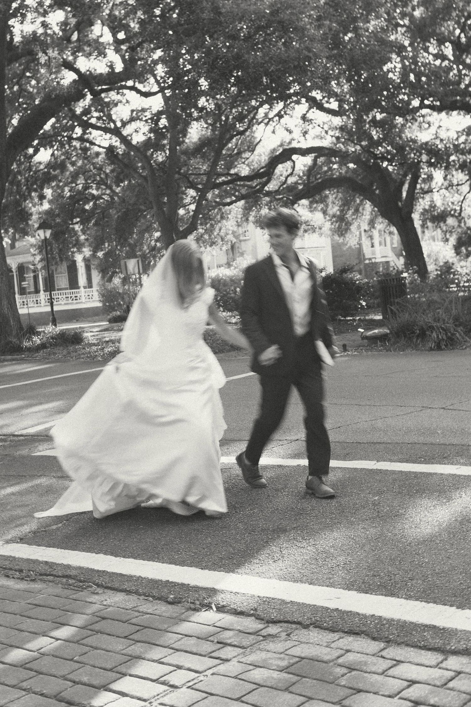 Black and white photo of bride and groom crossing a street lined with oak trees on their wedding day.
