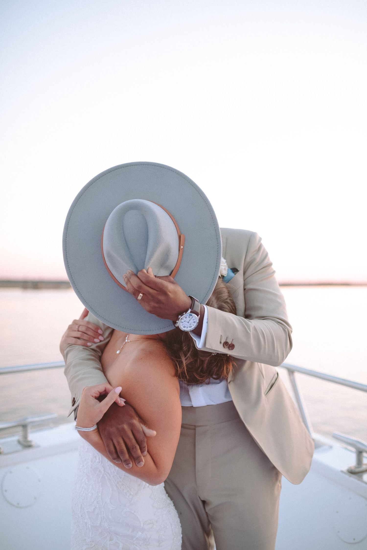 Newlyweds share a romantic kiss hidden behind a wide-brim gray hat on a yacht at sunset.