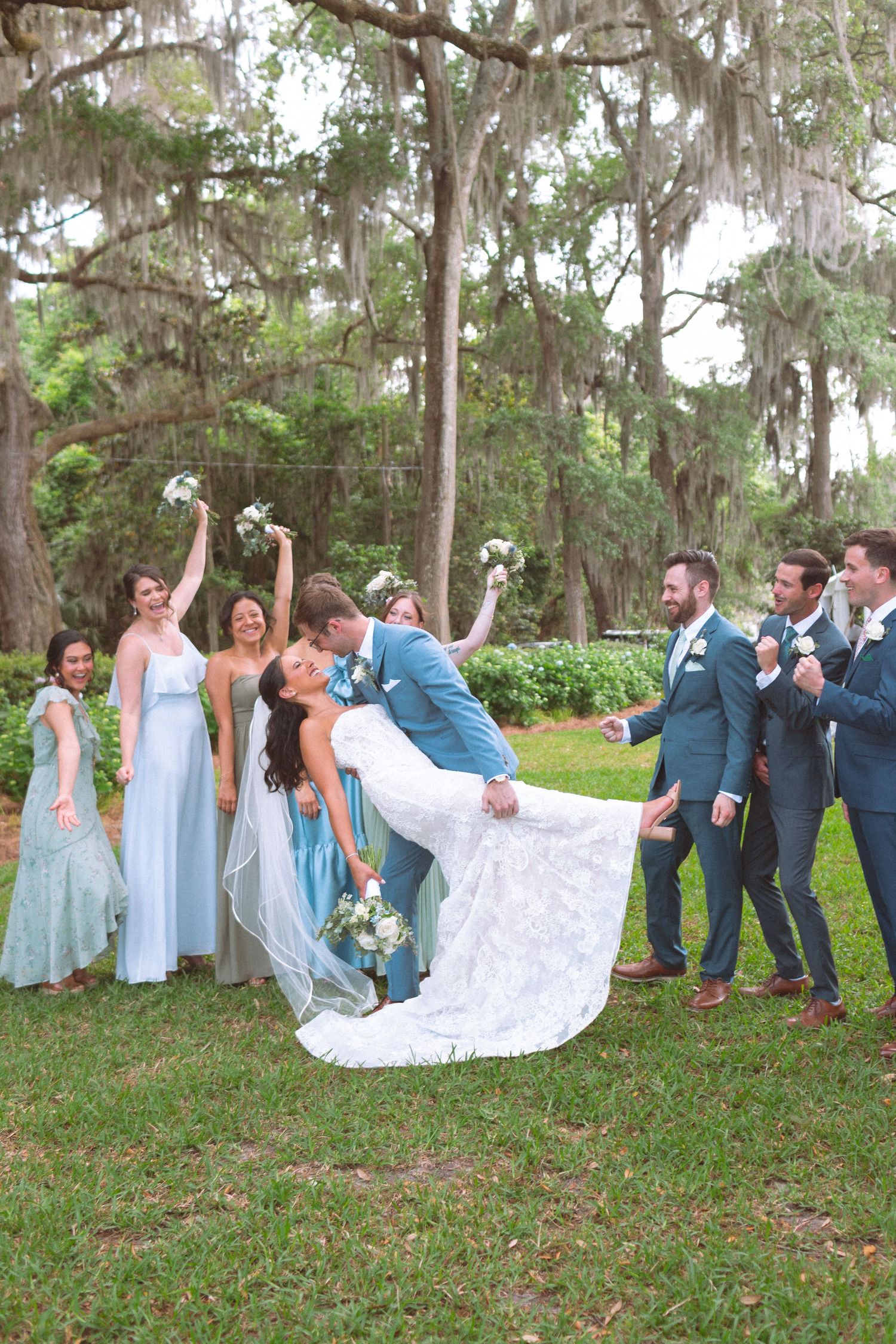 Groom dipping bride surrounded by cheering wedding party in blue attire under moss-covered trees.