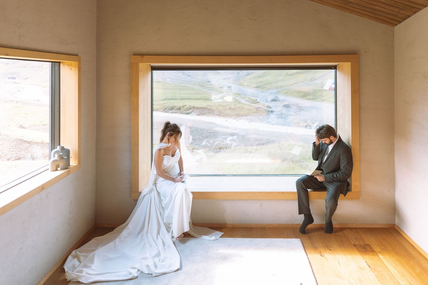 A bride in a white veil gown and groom sit apart in a modern room with a scenic mountain window.