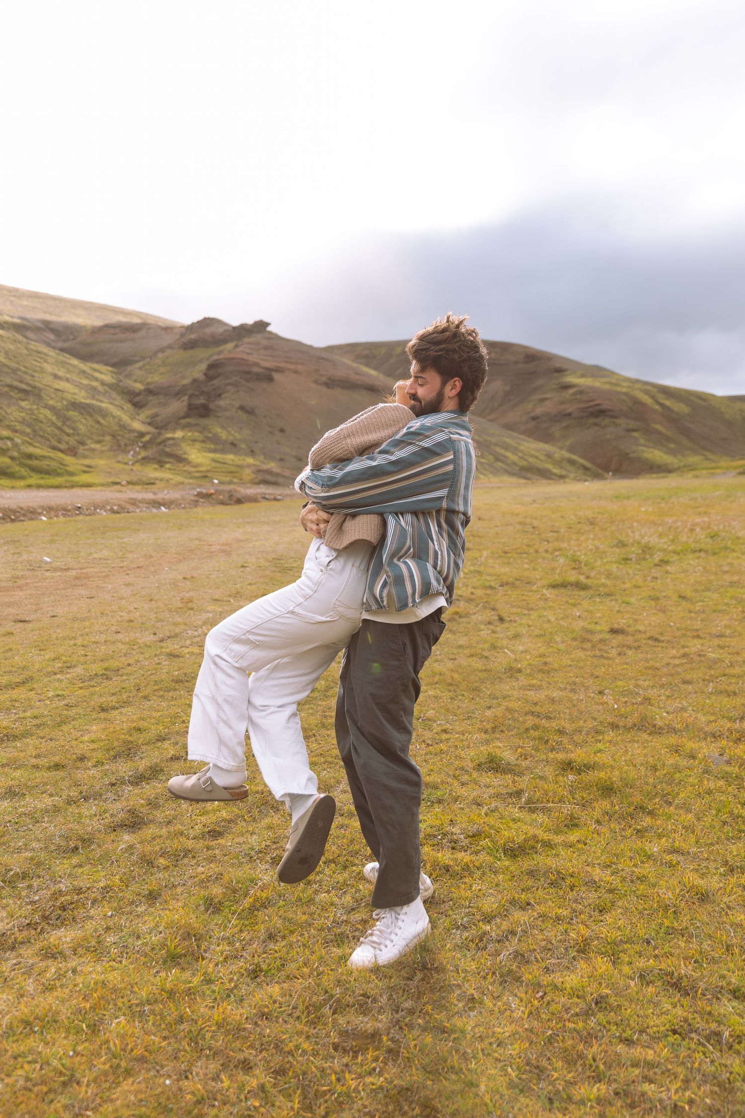 Man lifting woman joyfully in a grassy mountain field, both laughing and embracing playfully.