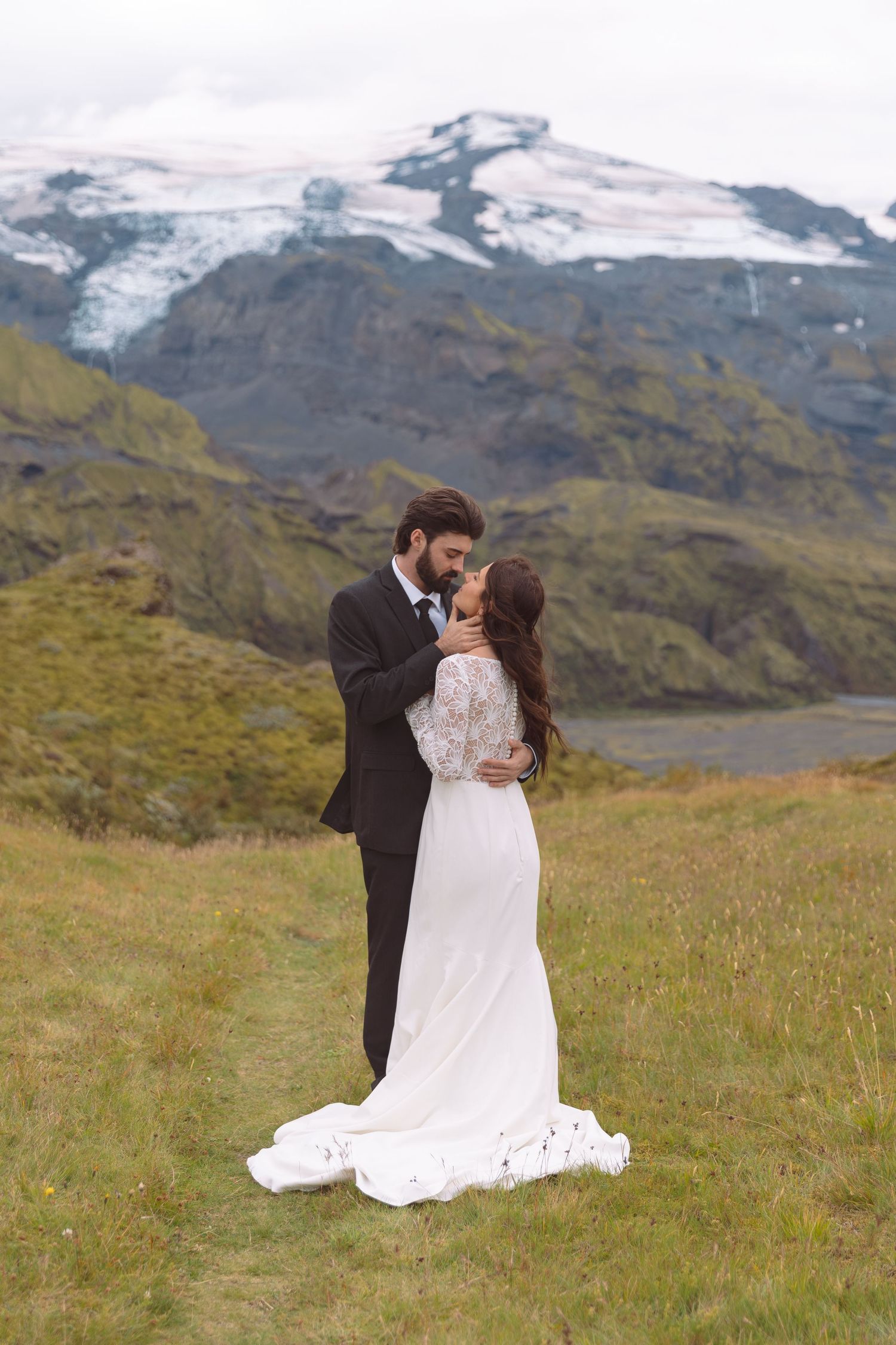 Groom tenderly kisses bride on forehead in a romantic full-length wedding portrait with glacier mountains.
