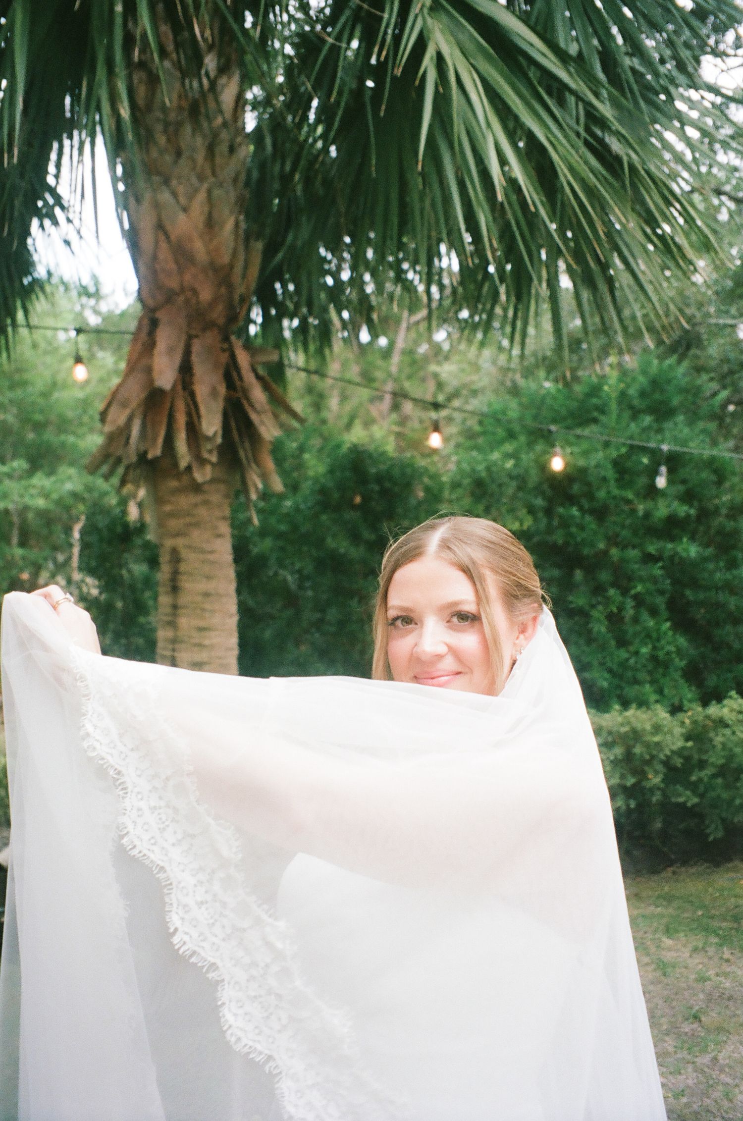 Smiling bride holding up her lace-trimmed veil outdoors with palm trees and string lights in the background.
